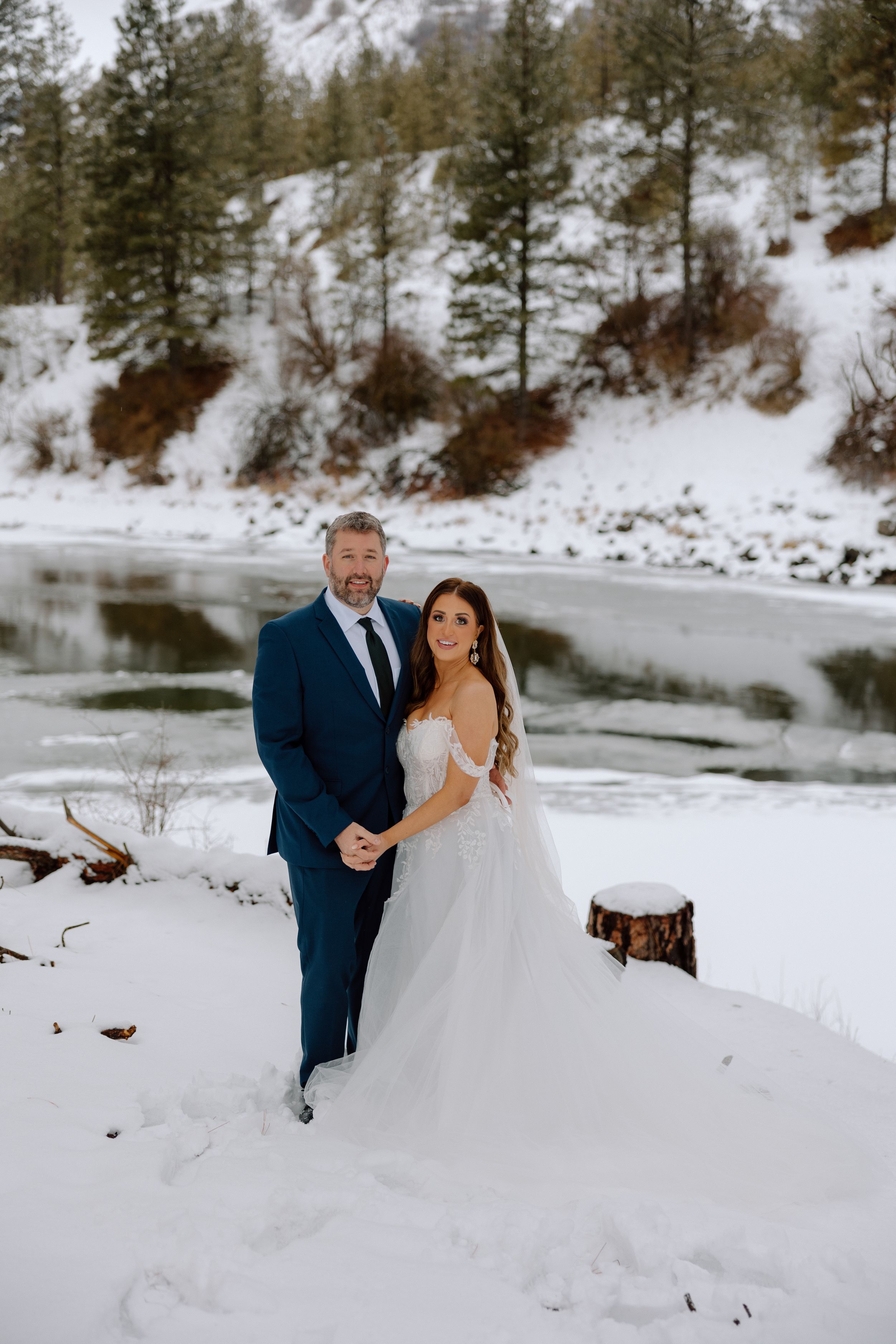 A winter wedding portrait captured by a frozen lake with snow-covered mountains in the background.