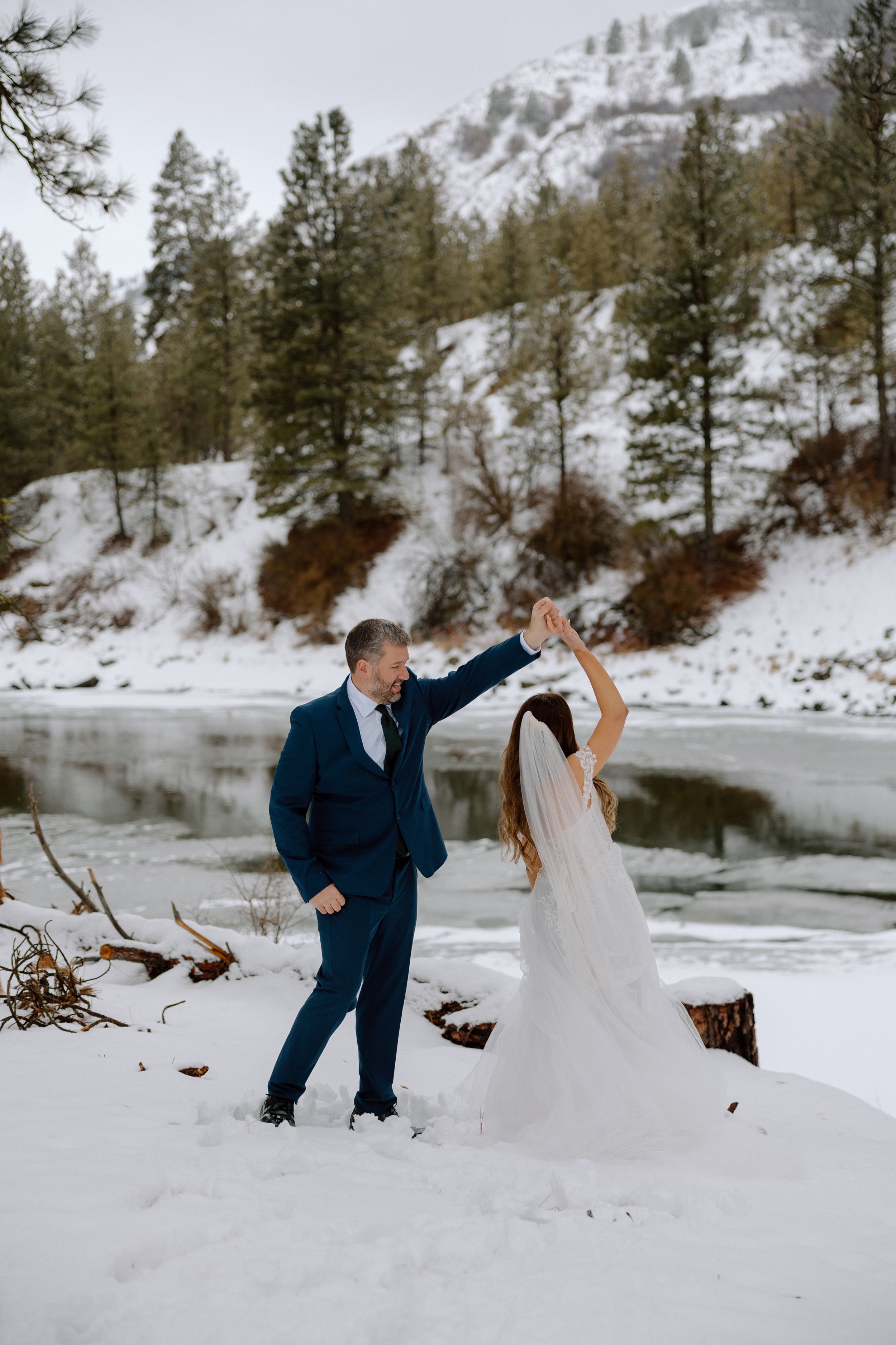 A couple dances joyfully in the snow with a scenic mountain backdrop and frozen lake.
