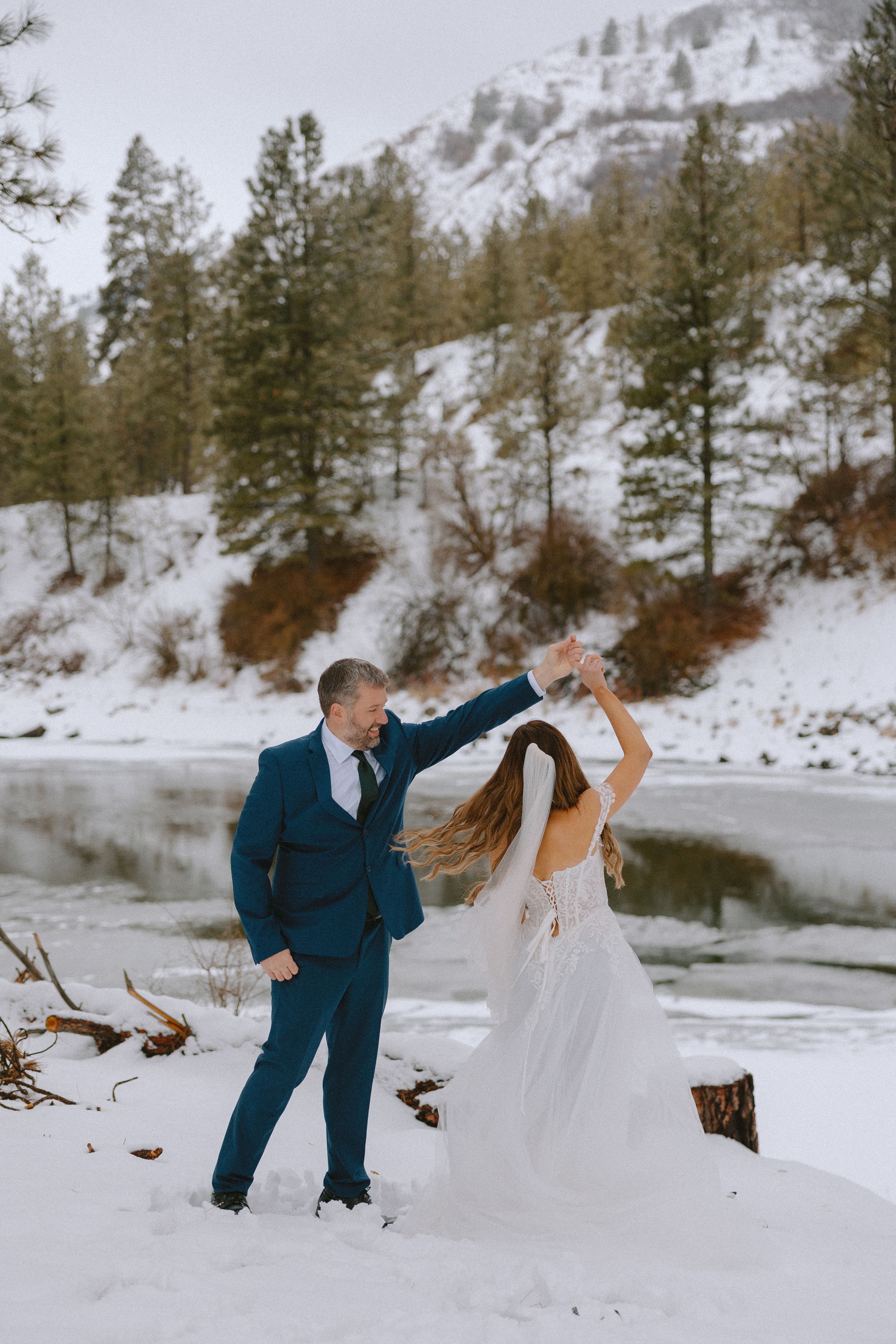 A couple shares a dance in the snow beside a frozen lake with snow-capped mountains in the distance.