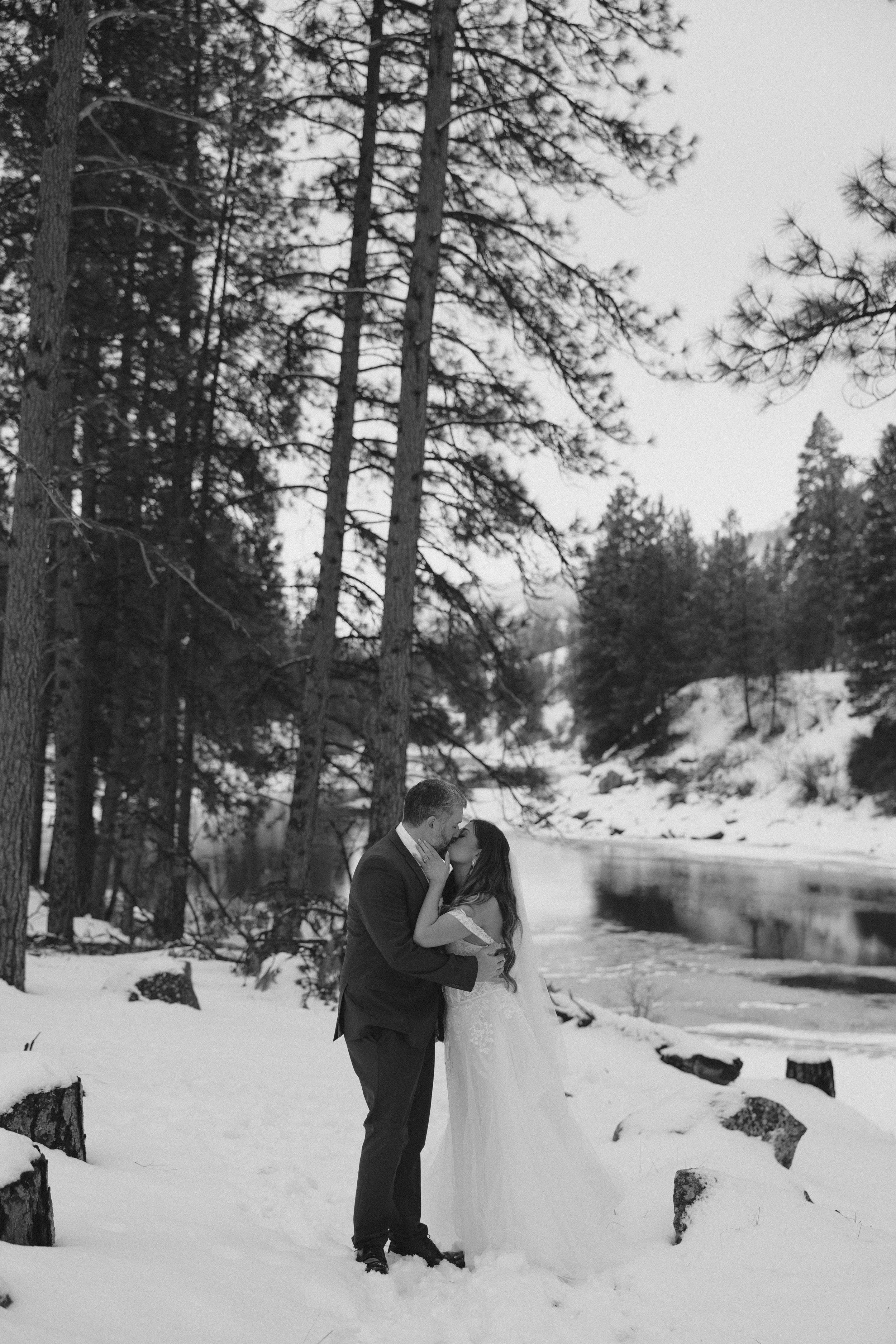 A romantic winter wedding photo of a couple embracing by a snowy riverside surrounded by tall pine trees.