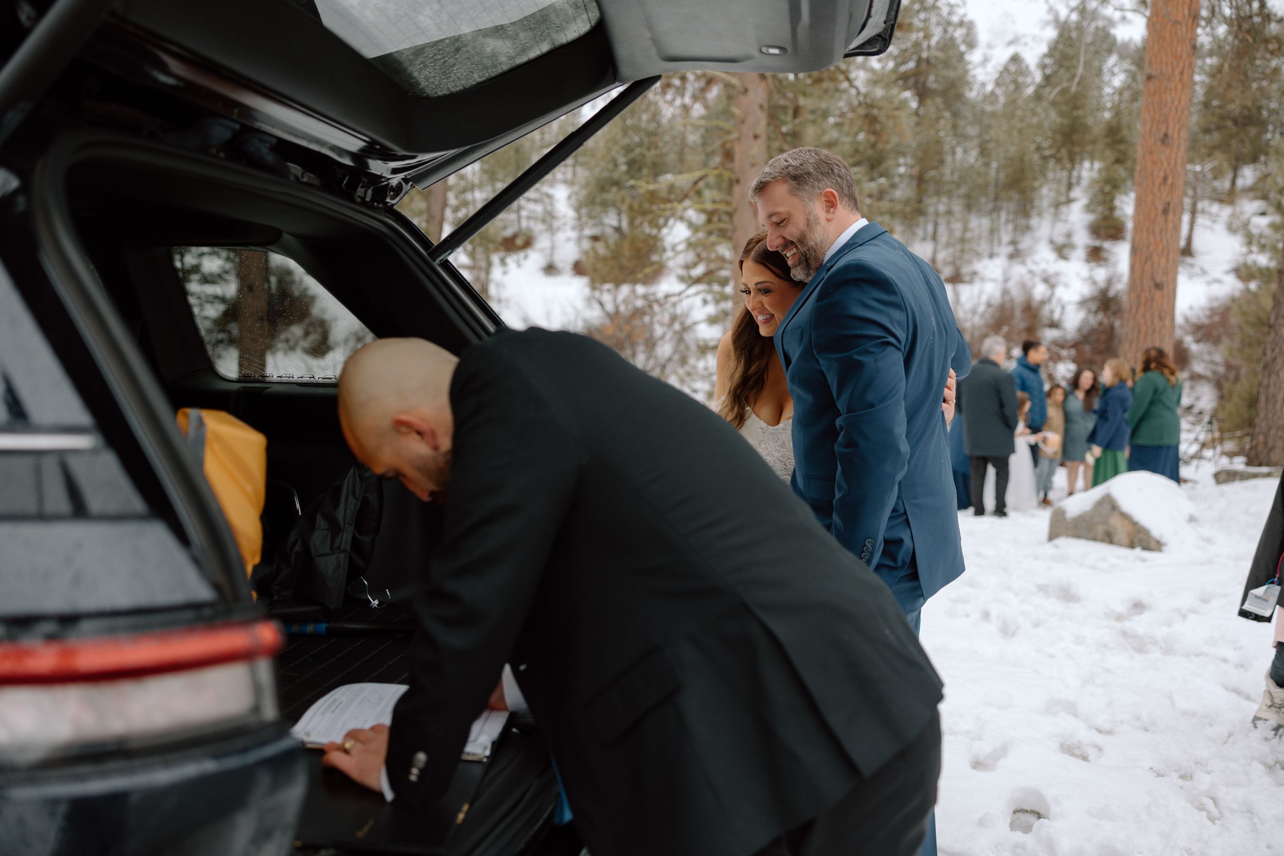 People gather around a car trunk in a snowy landscape during a winter wedding event.
