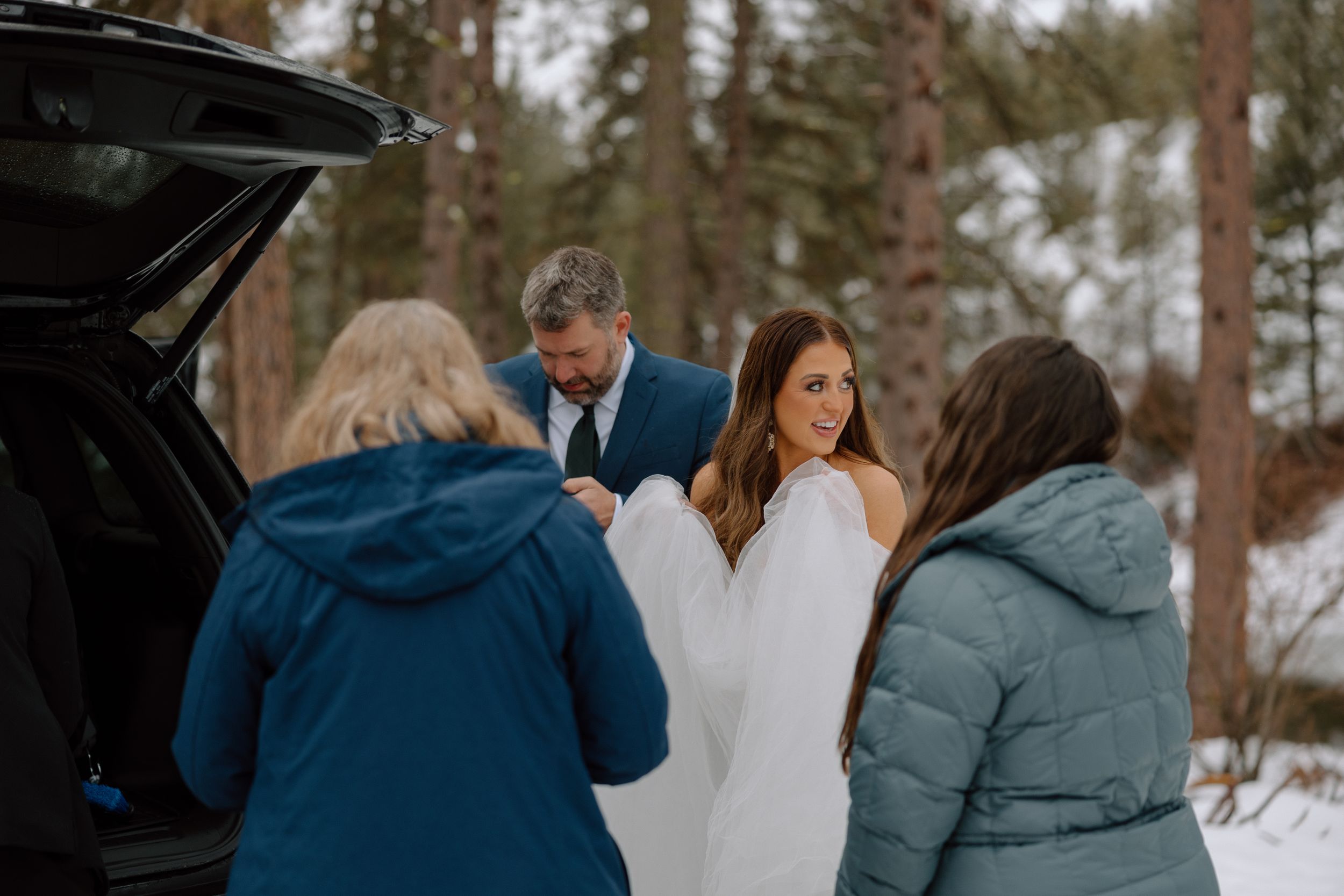 Wedding party gathers around a car in a snowy forest setting during a winter celebration.