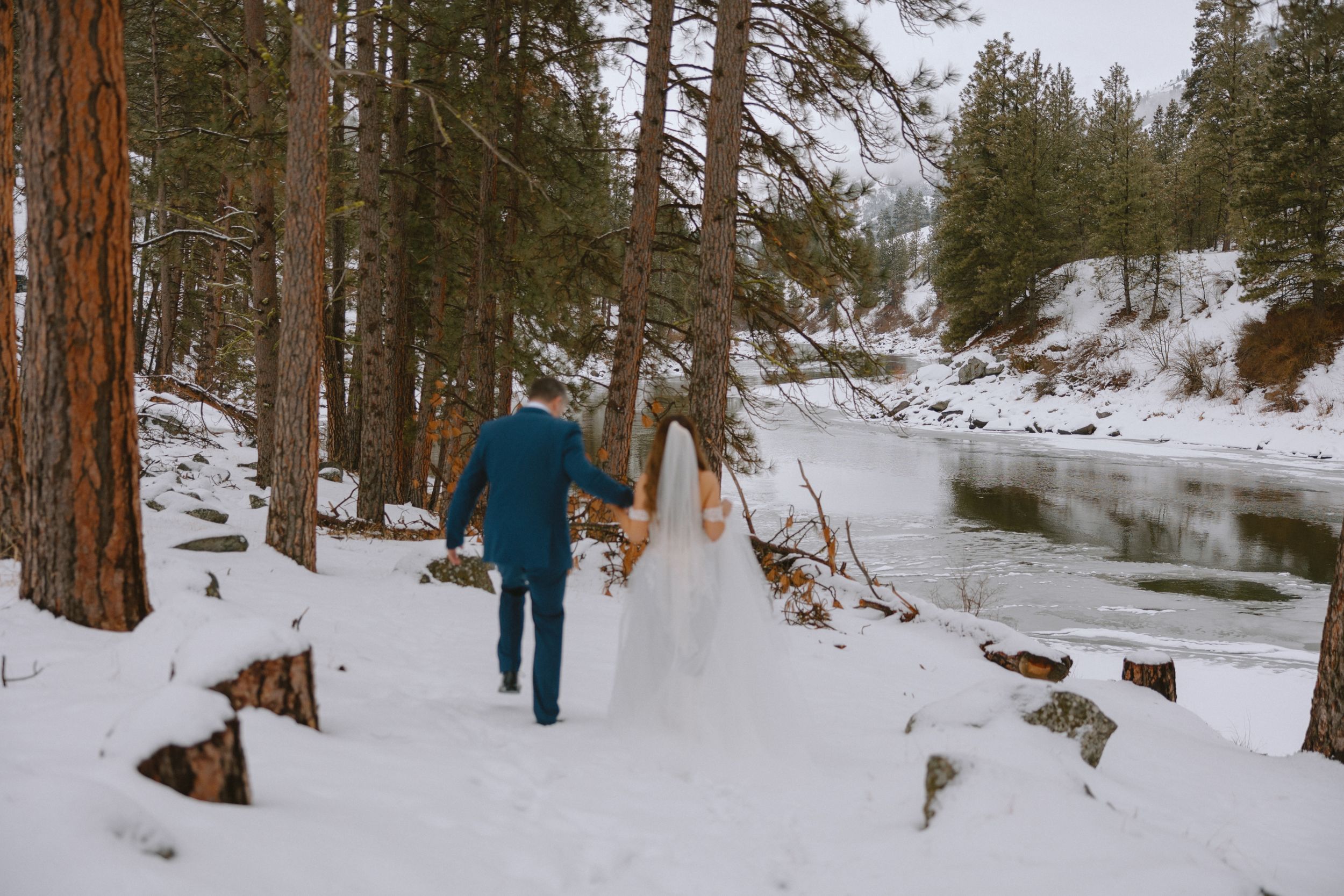 A couple walks hand in hand through a snowy forest path alongside a partially frozen river.