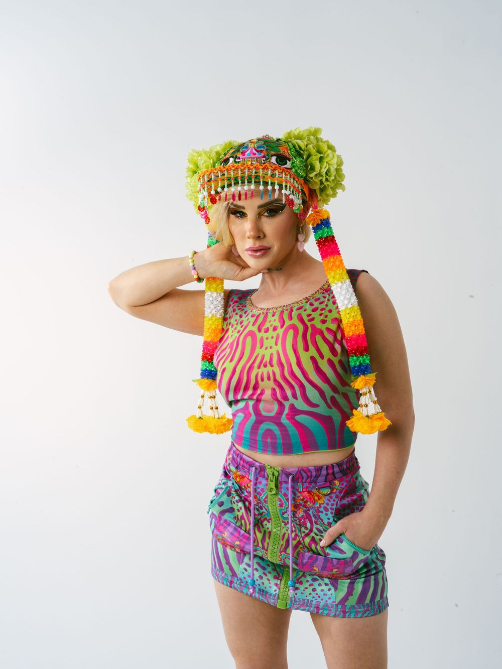Colorful festival outfit with bright patterns, featuring a floral headpiece and tasseled accessories against white background.