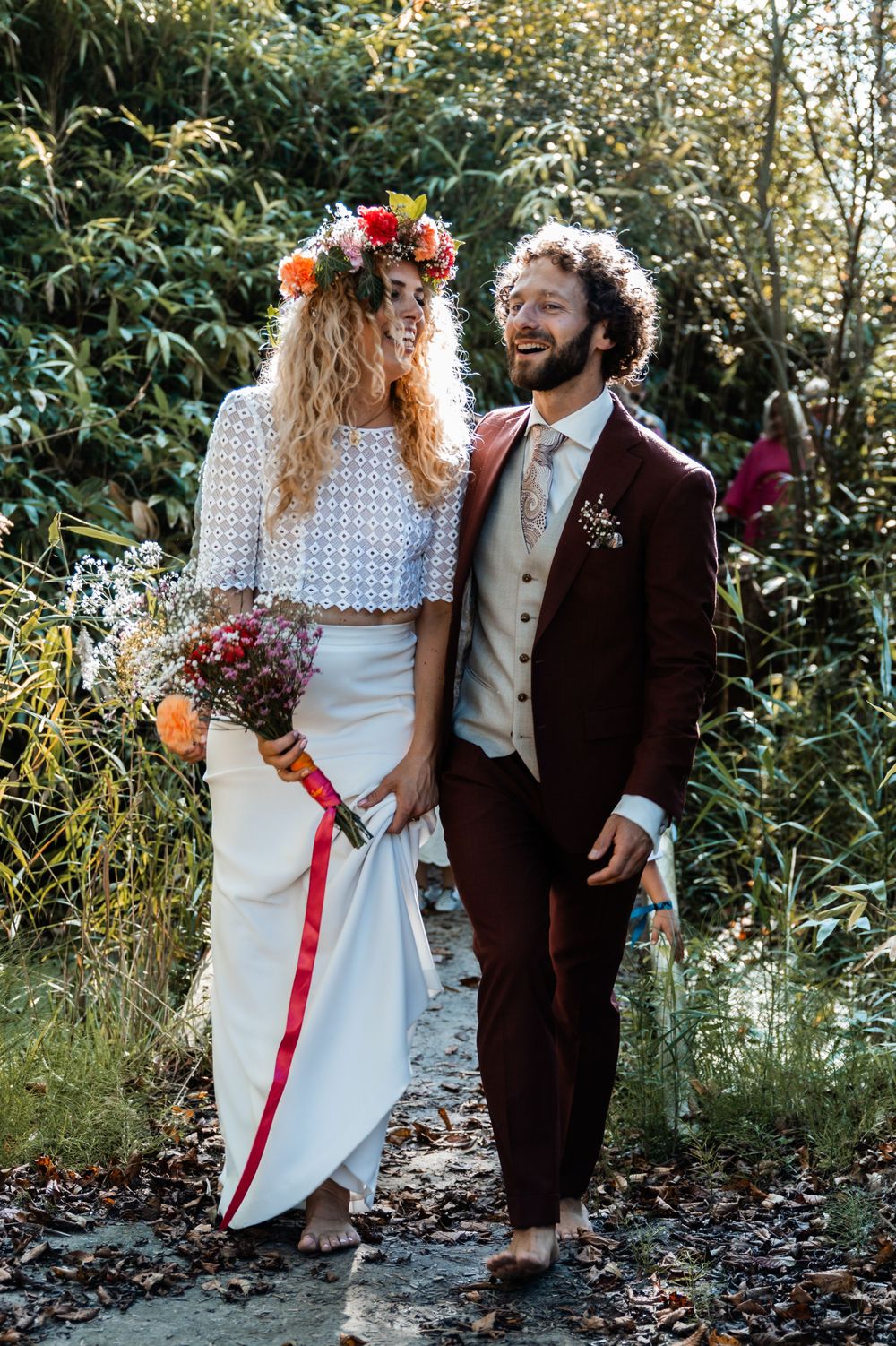 Newlyweds share a joyful moment in nature, with bride wearing floral crown and two-piece dress and groom in burgundy suit.