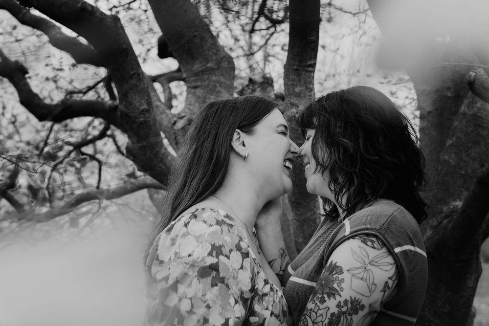 Black and white photo of two people in floral dresses sharing a joyful moment under a tree.