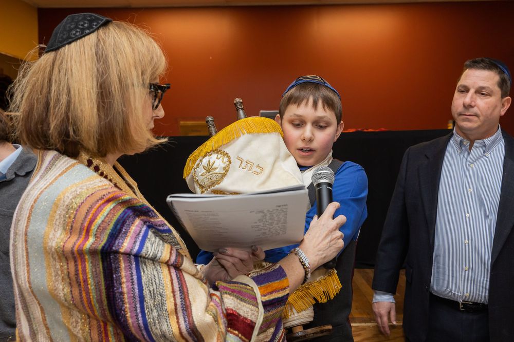 Young person wearing a kippah reads from a document during what appears to be a bar mitzvah ceremony.