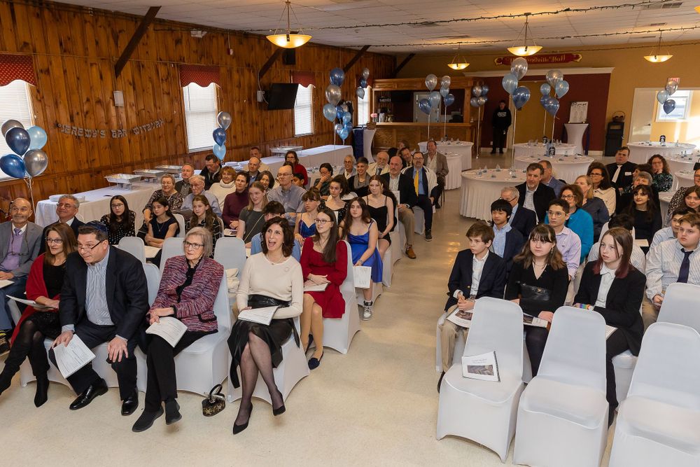 Large audience seated in rows at a community event with wooden paneled walls and blue balloon decorations.