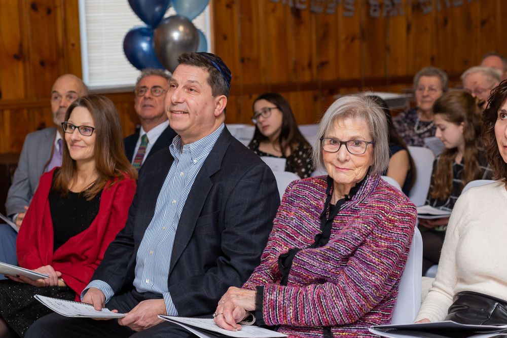 Attendees seated in rows at formal ceremony with blue balloons and wooden interior decor.