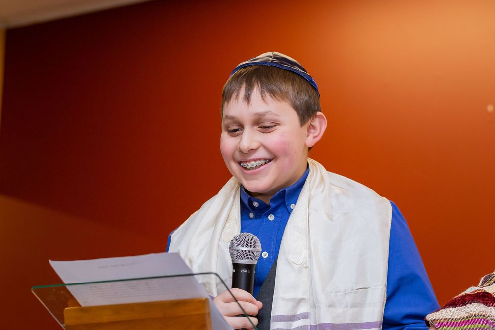 Young person in white and blue attire smiles while speaking into microphone against orange wall.