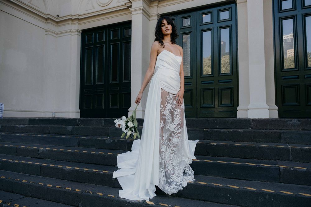 A stunning white wedding gown with lace details and flowing train stands on grand stone steps against dark green doors.