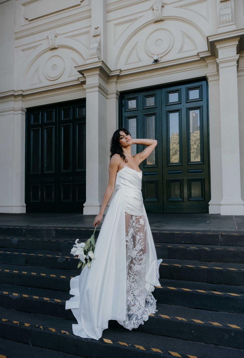 A bride in a flowing white gown with lace details poses elegantly on dark steps against vintage dark green doors.