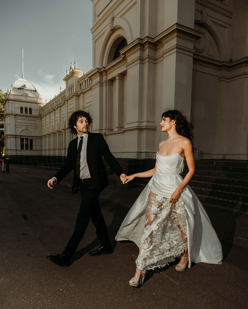 A couple in wedding attire runs playfully along a historic building exterior with grand architecture and arched windows.