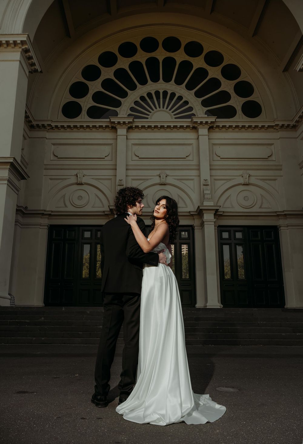 A couple in wedding attire embraces under an ornate archway with a decorative fanlight window above.