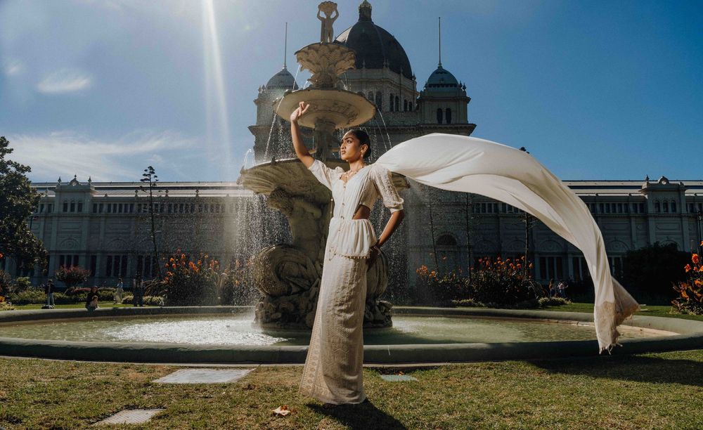 A romantic wedding couple shares an intimate moment in front of a grand palace with dramatic flowing veil in the sunlight.