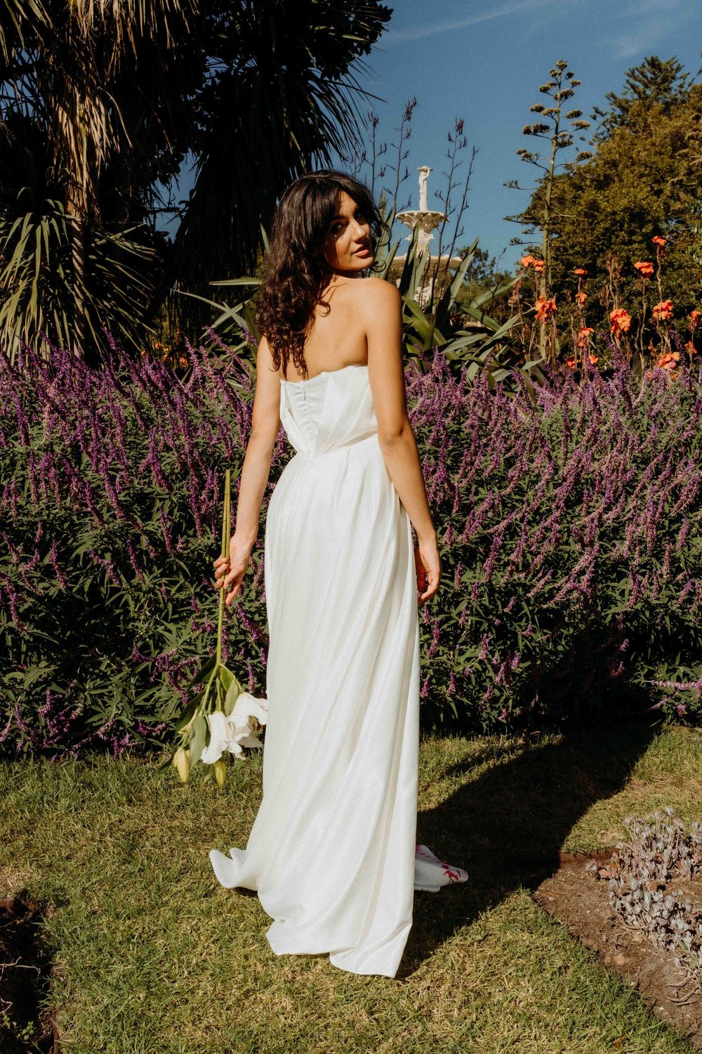 Elegant white wedding dress against a backdrop of vibrant purple flowers and palm trees in a garden setting.