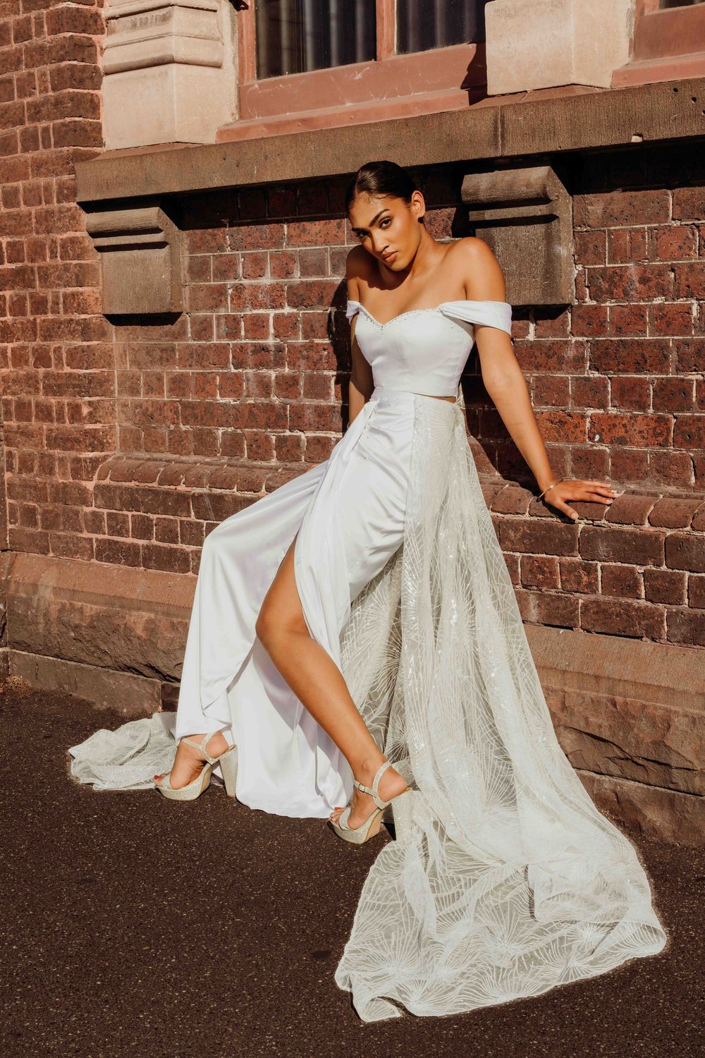 A stunning off-shoulder wedding gown with leg slit shown in an editorial pose against a rustic brick wall in warm sunlight.