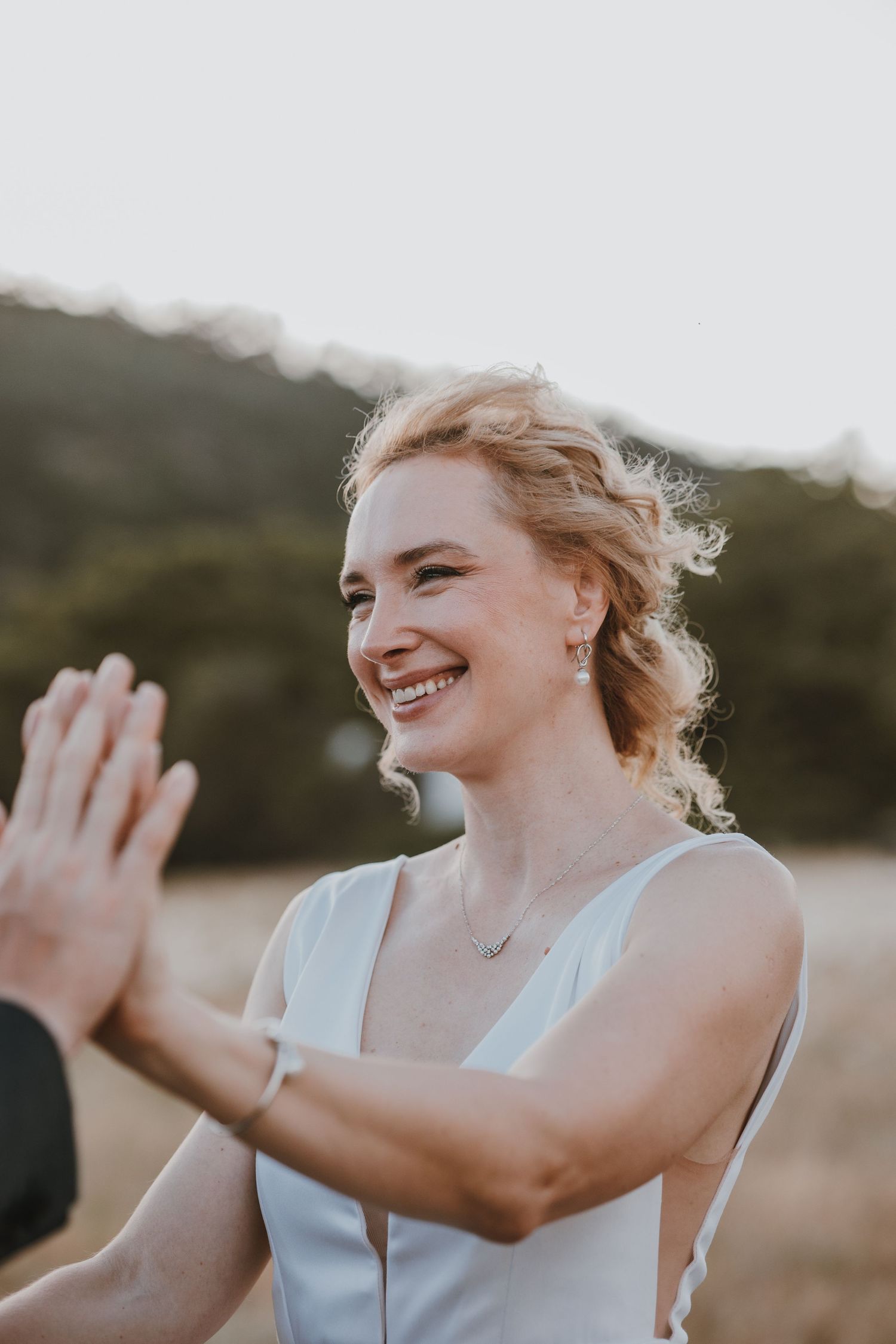 A joyful bride in a white sleeveless dress laughs while outdoors during golden hour.