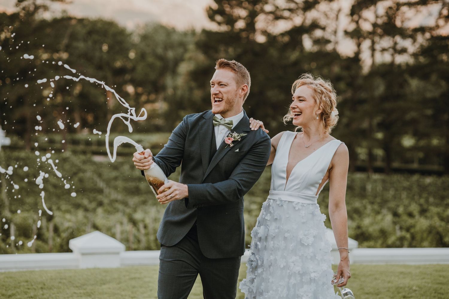Wedding couple celebrates with champagne spray in outdoor garden setting during golden hour.