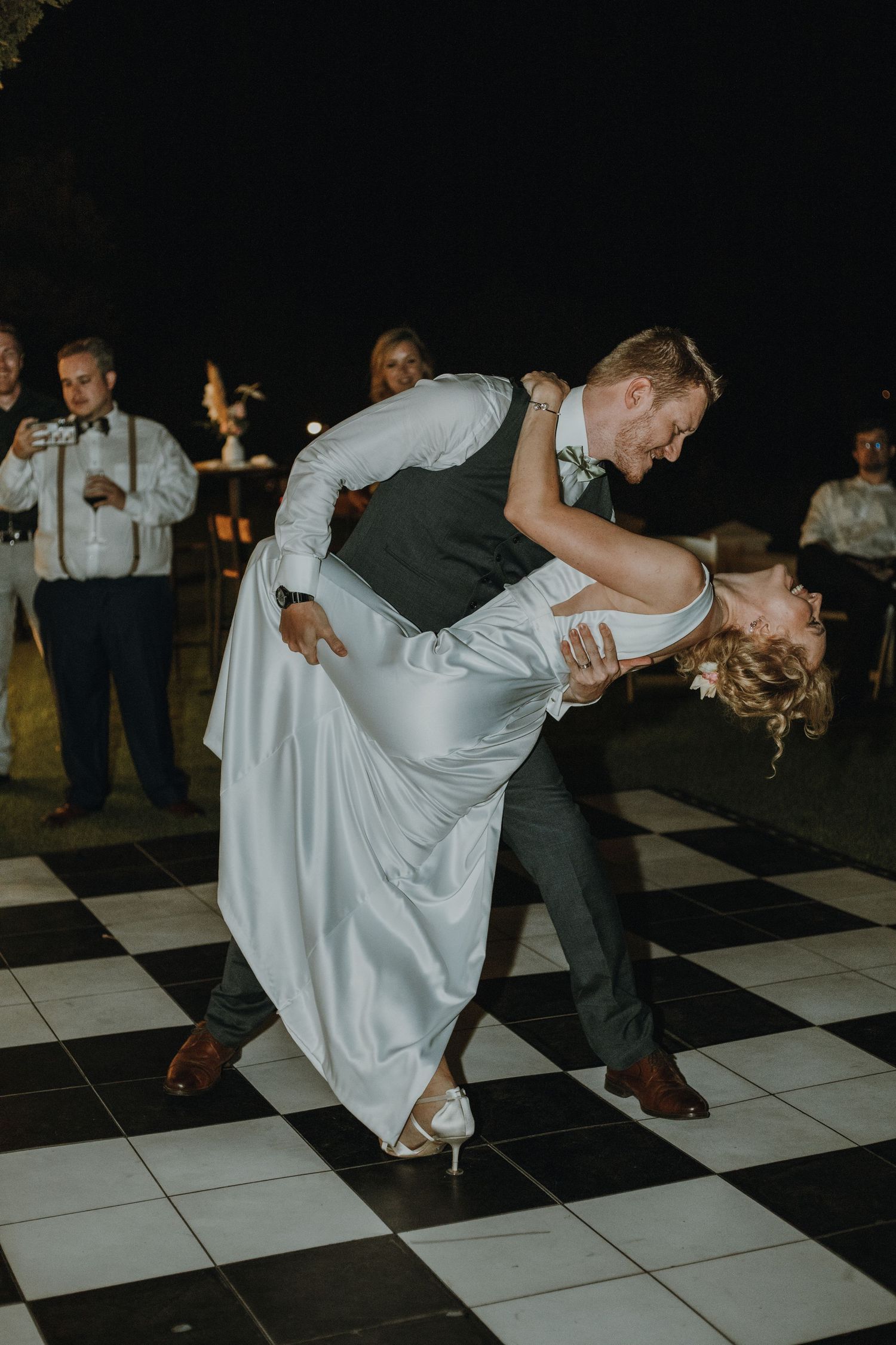 A couple performs an elegant dip during their first dance on a black and white checkered dance floor.