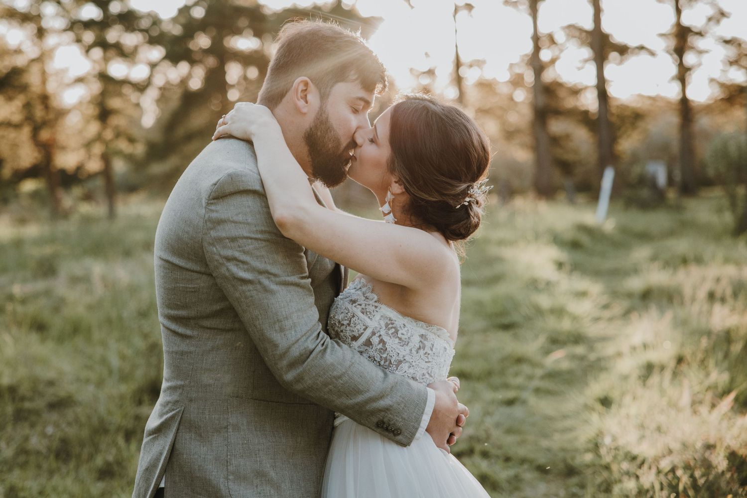 A romantic couple embrace and kiss in an outdoor field at golden hour while wearing wedding attire.