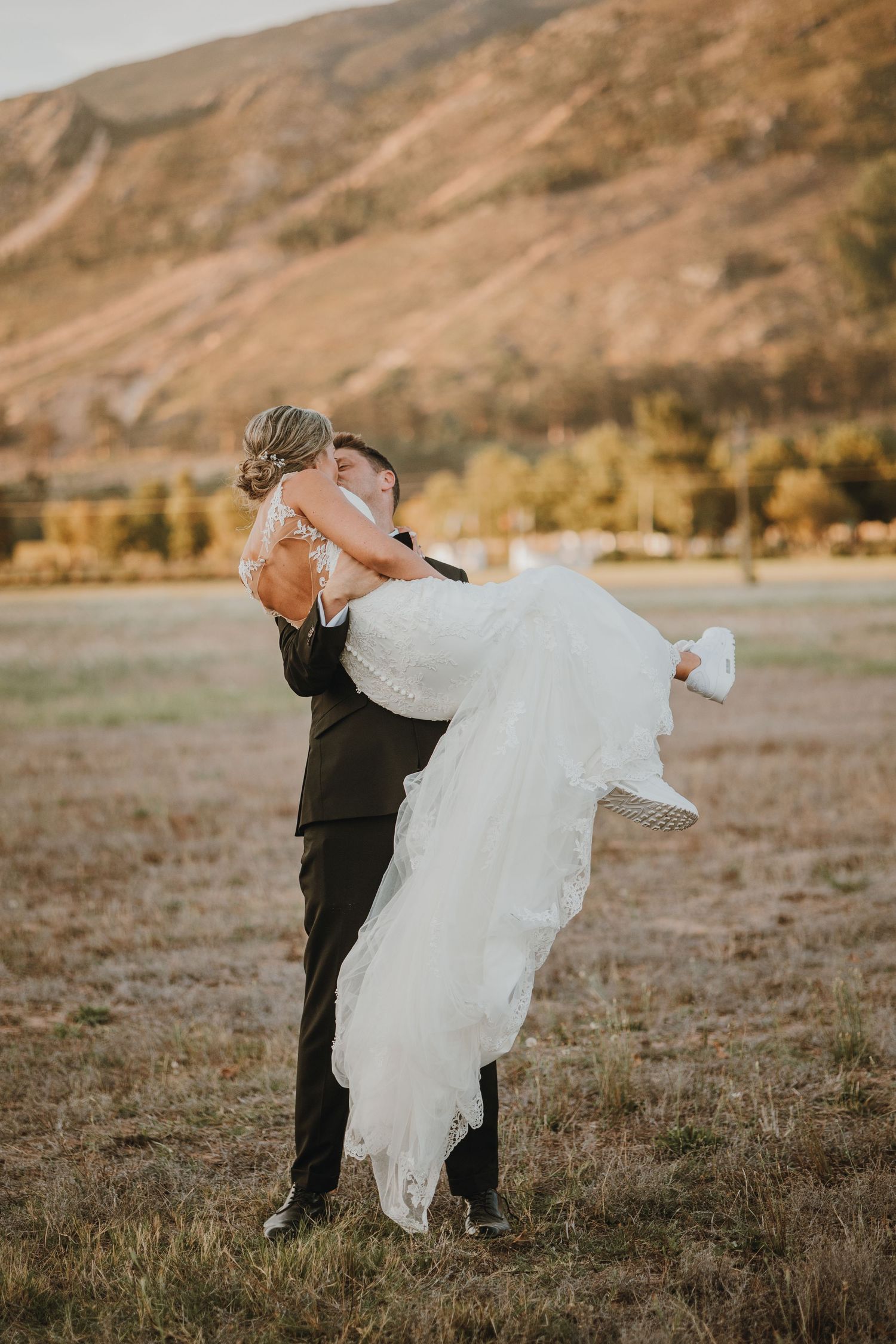 A couple in formal wedding attire share an intimate moment in a field with golden mountains in the background.