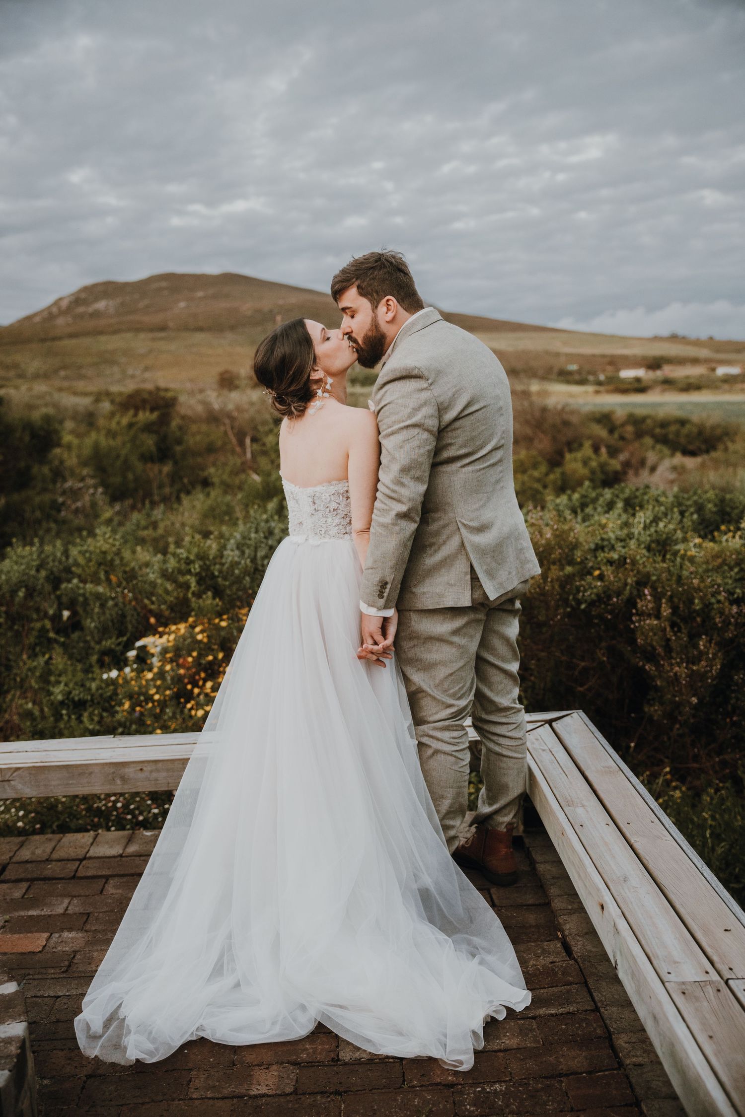 A couple shares a romantic kiss on a wooden deck overlooking rolling hills on their moody wedding day.