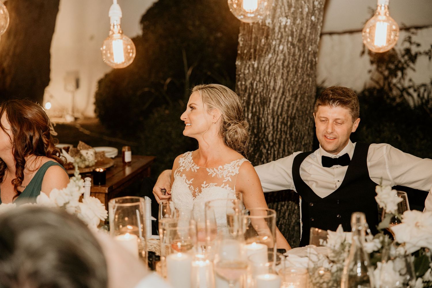 Bride and groom share a candid moment at an outdoor evening wedding reception with warm lighting and rustic decor.