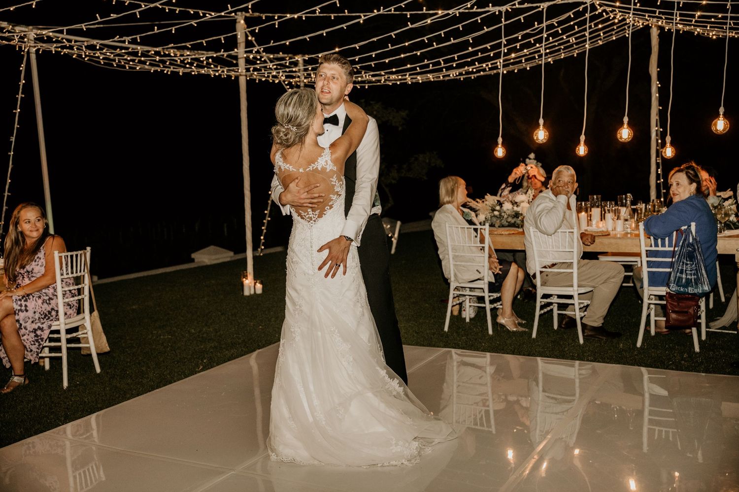 A couple shares a romantic dance on an outdoor dance floor under string lights with wedding reception tables in the background.