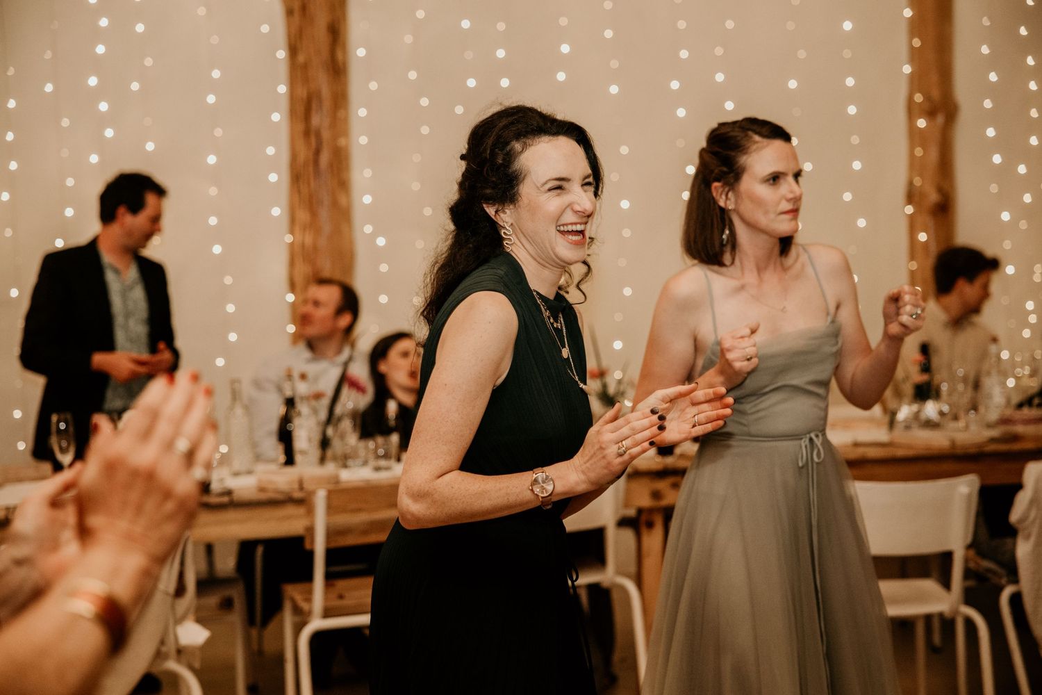 Wedding reception guests in formal attire dancing and celebrating with string lights in rustic wooden venue setting.