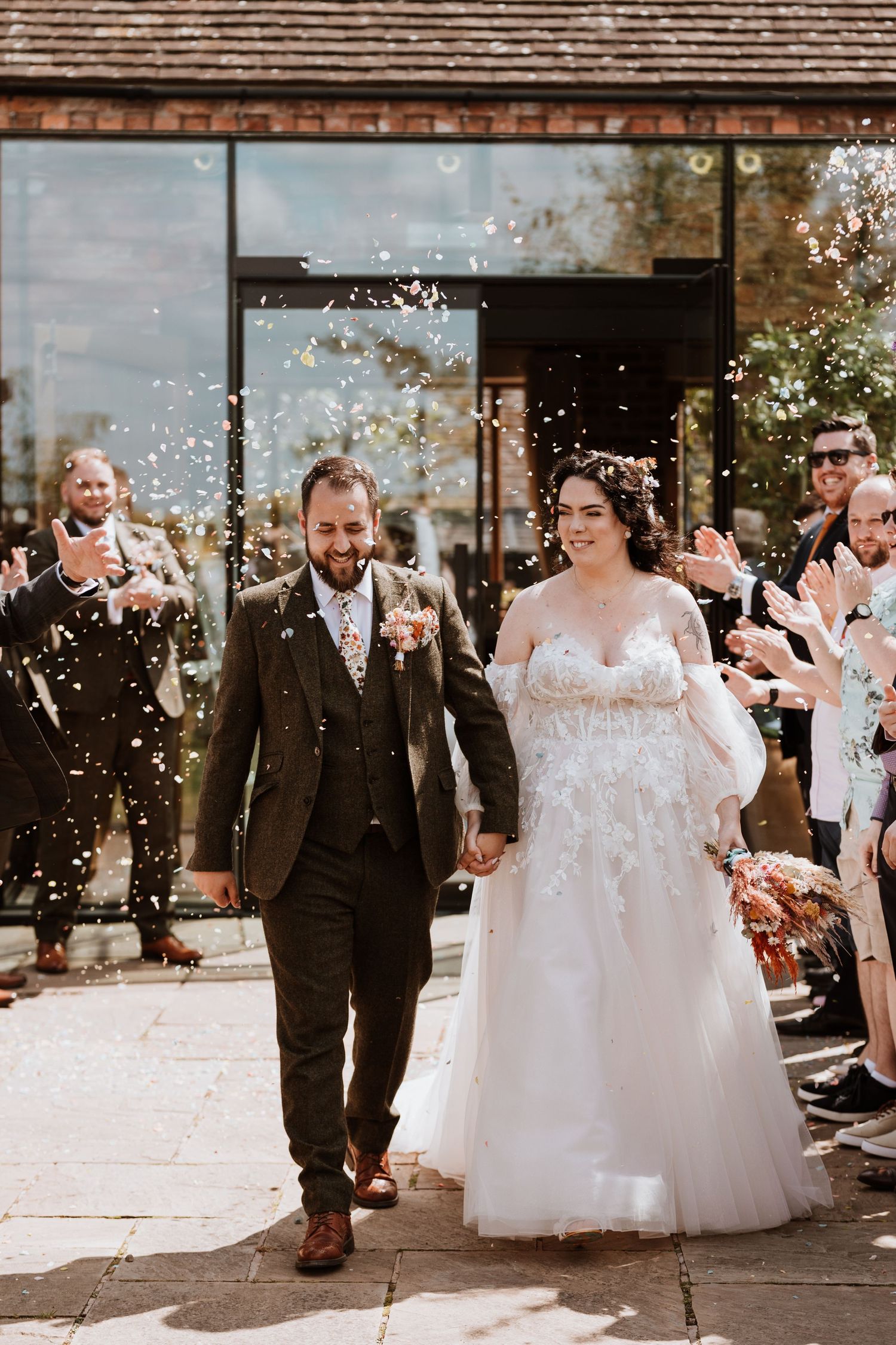 Newlyweds walk down stone pathway while guests shower them with confetti outside at Curradine barns.