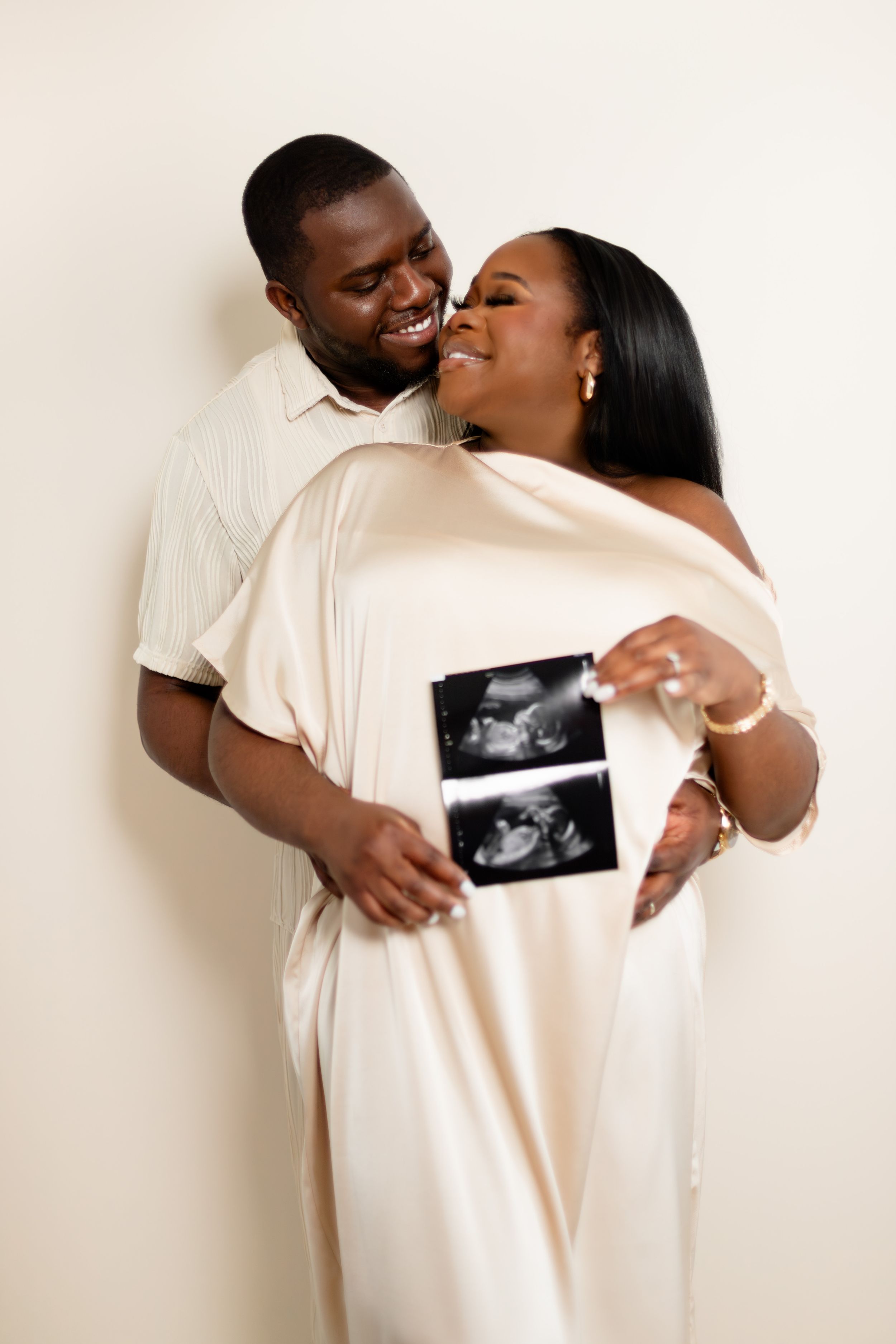 A loving couple shares a joyful moment holding ultrasound photos while embracing against a light neutral background.
