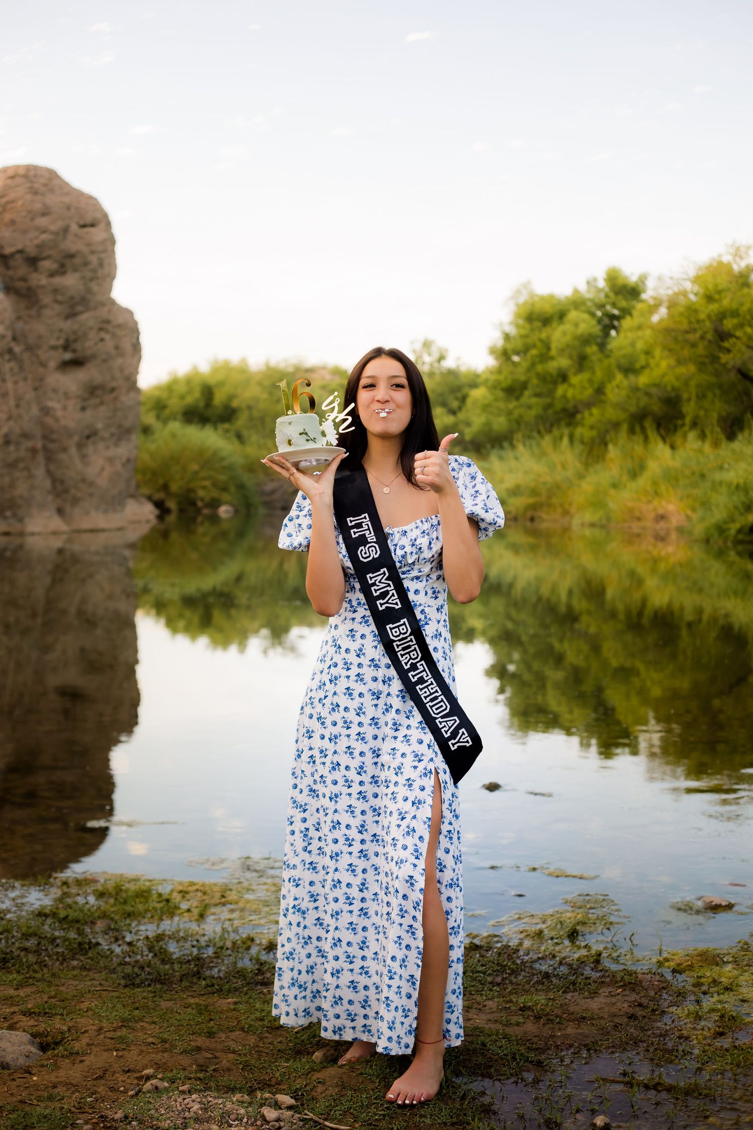 Portrait of a teenage girl with a sweet sixteen birthday cake in a beautiful gown standing in a lush green river location in Arizona.