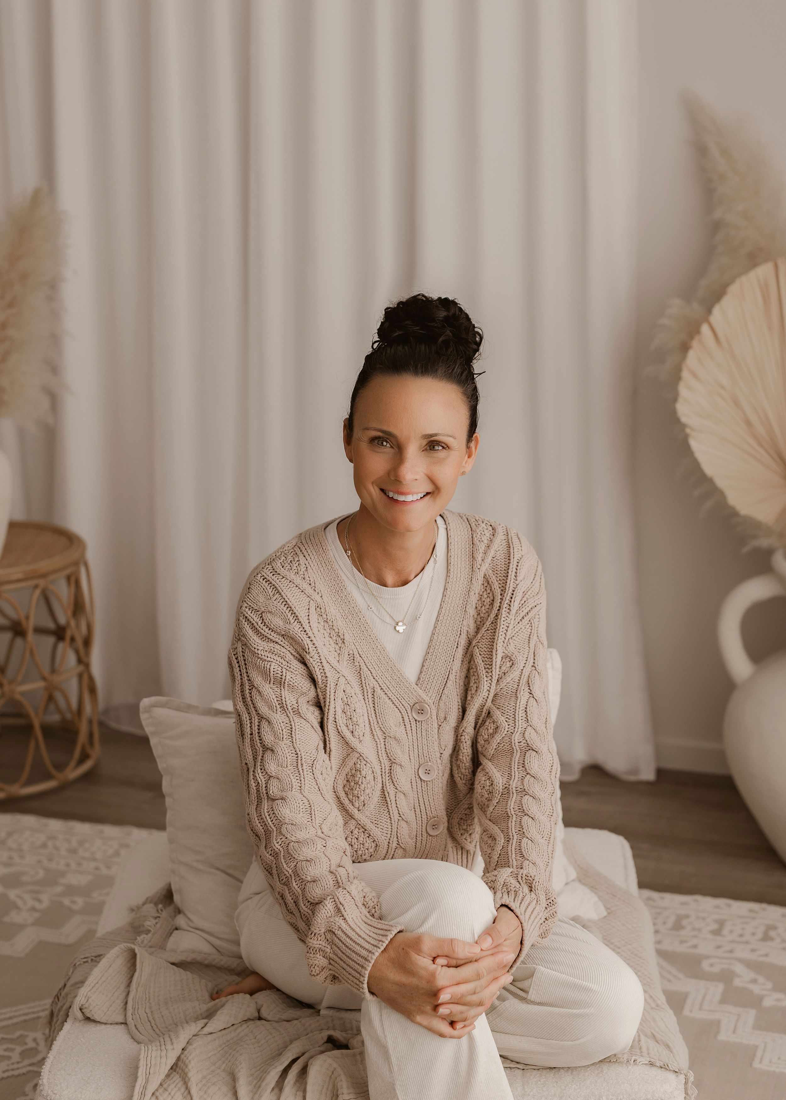 Person sits cross-legged wearing beige cable knit cardigan and white pants in cozy neutral room with dried palm decor.