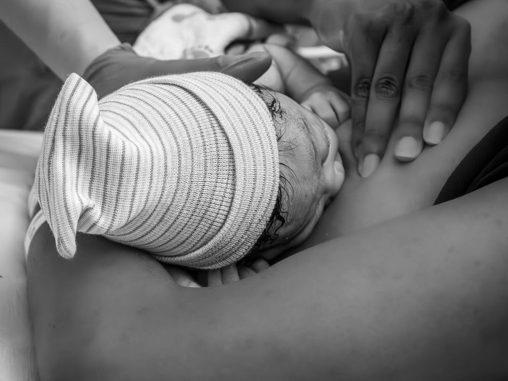 Newborn baby swaddled in a striped hospital cap rests against mother's chest in black and white.