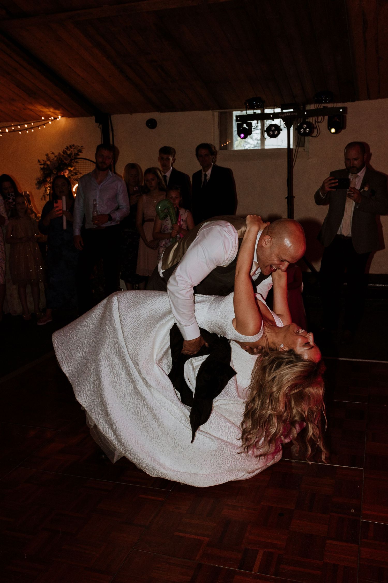 A couple performs a dramatic dip while dancing at a wedding reception with string lights and wood paneling visible at Stanford Farm.
