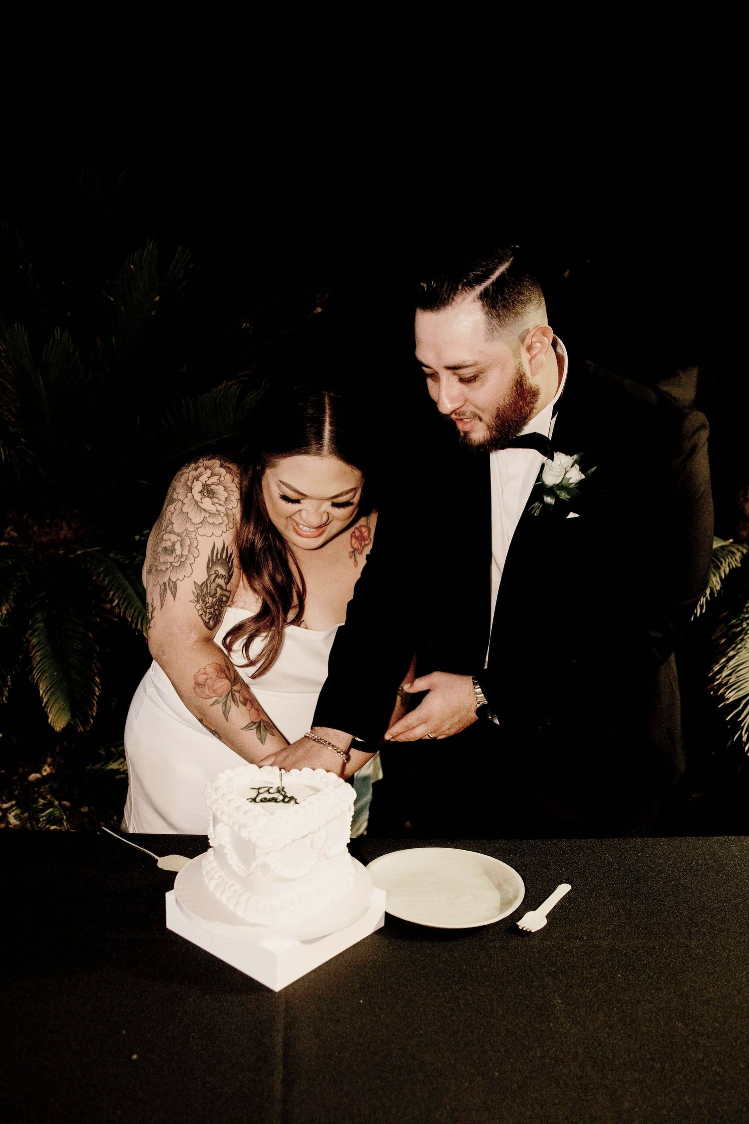 A couple in wedding attire laughs together while cutting their wedding cake at a nighttime reception.