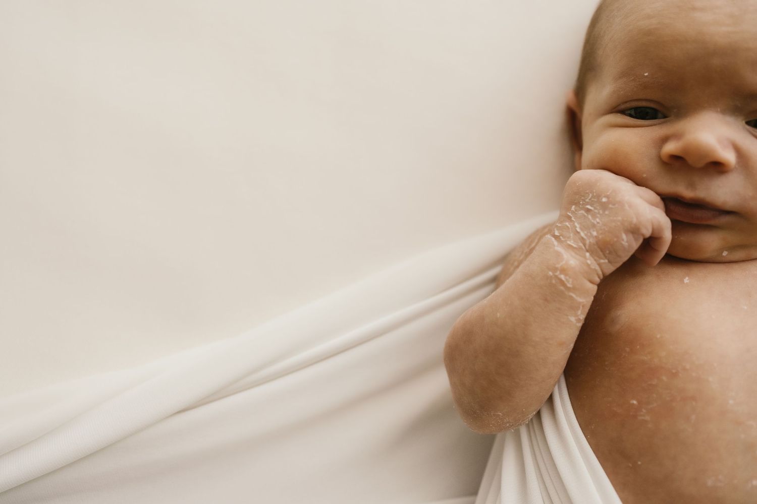 Infant in white clothes curiously sucking on their finger while lying on a light beige surface.