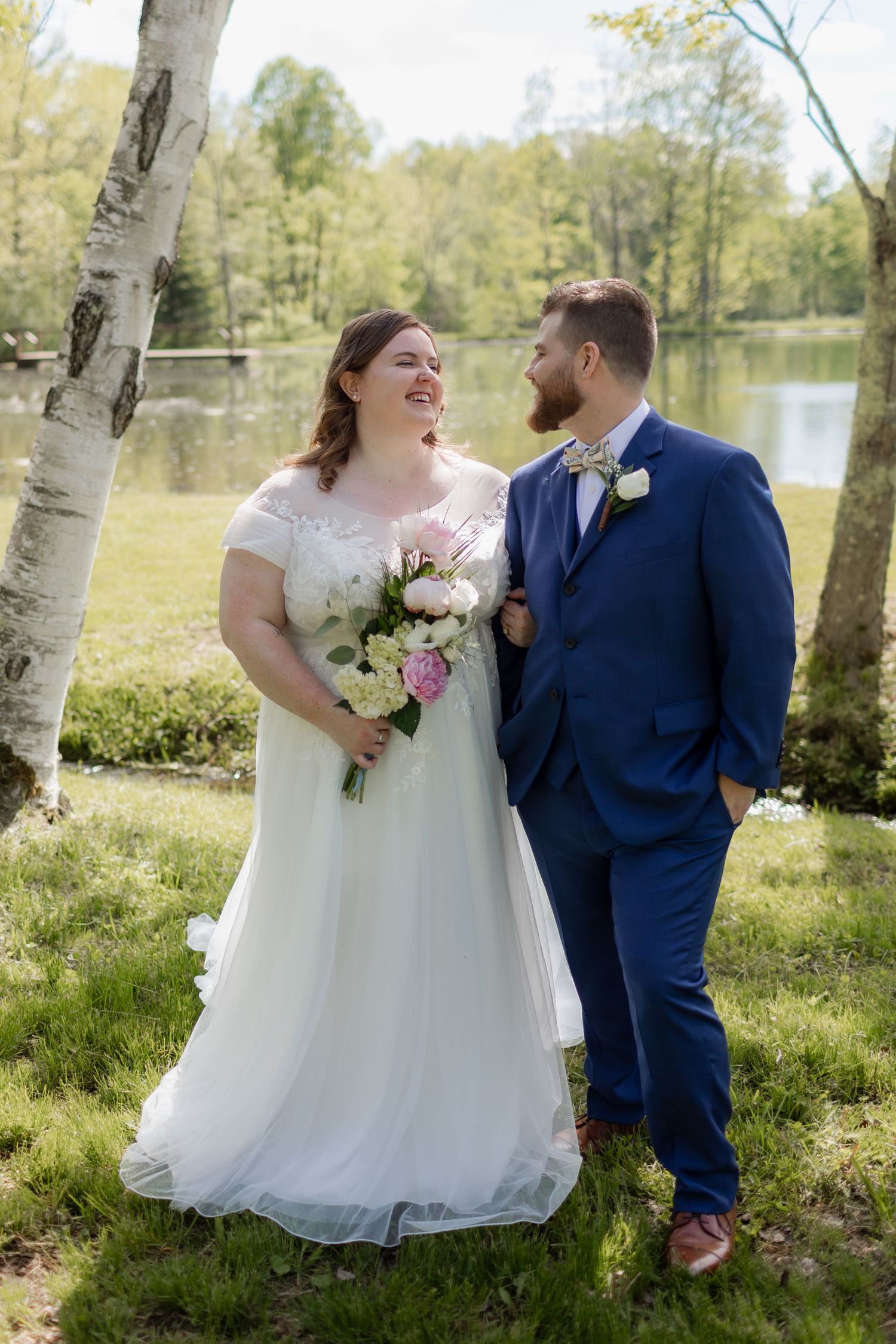 A couple shares a joyful candid moment by a lake in springtime, with the bride holding pink and white flowers.