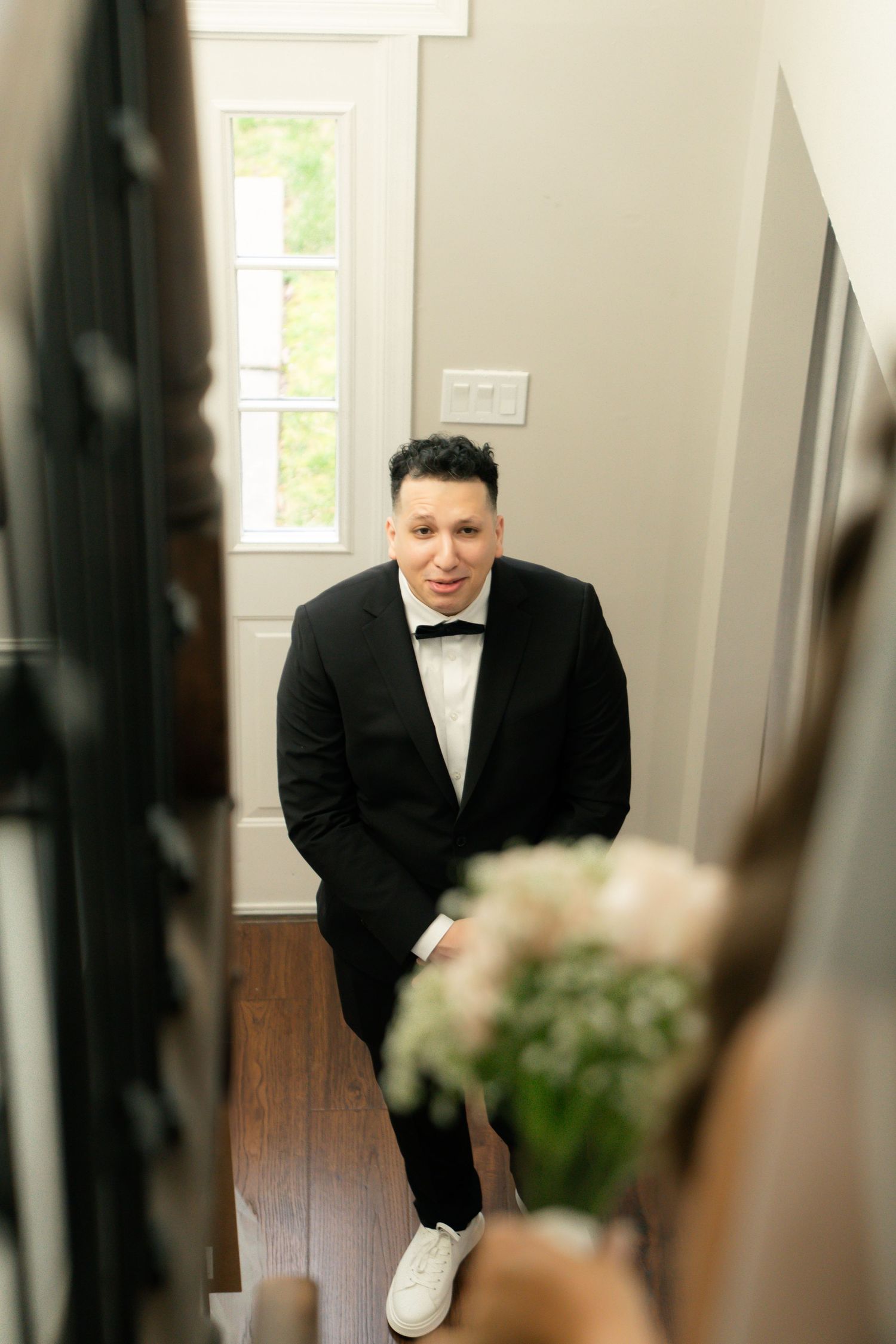 A person in a formal black tuxedo and bow tie stands in a hallway wearing white sneakers for contrast.