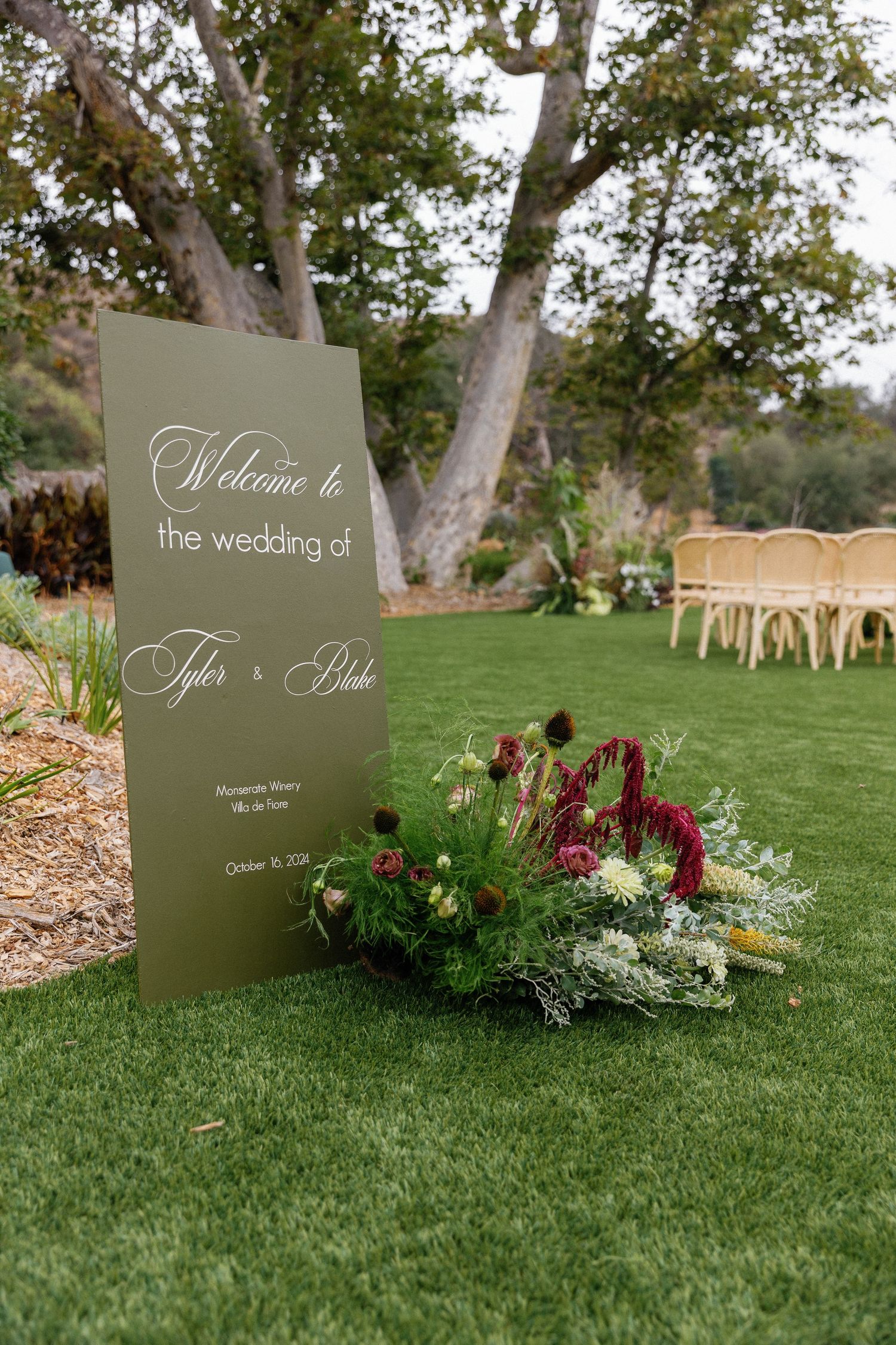 Elegant sage green wedding welcome sign with burgundy floral arrangement sits on manicured lawn with chairs in background.