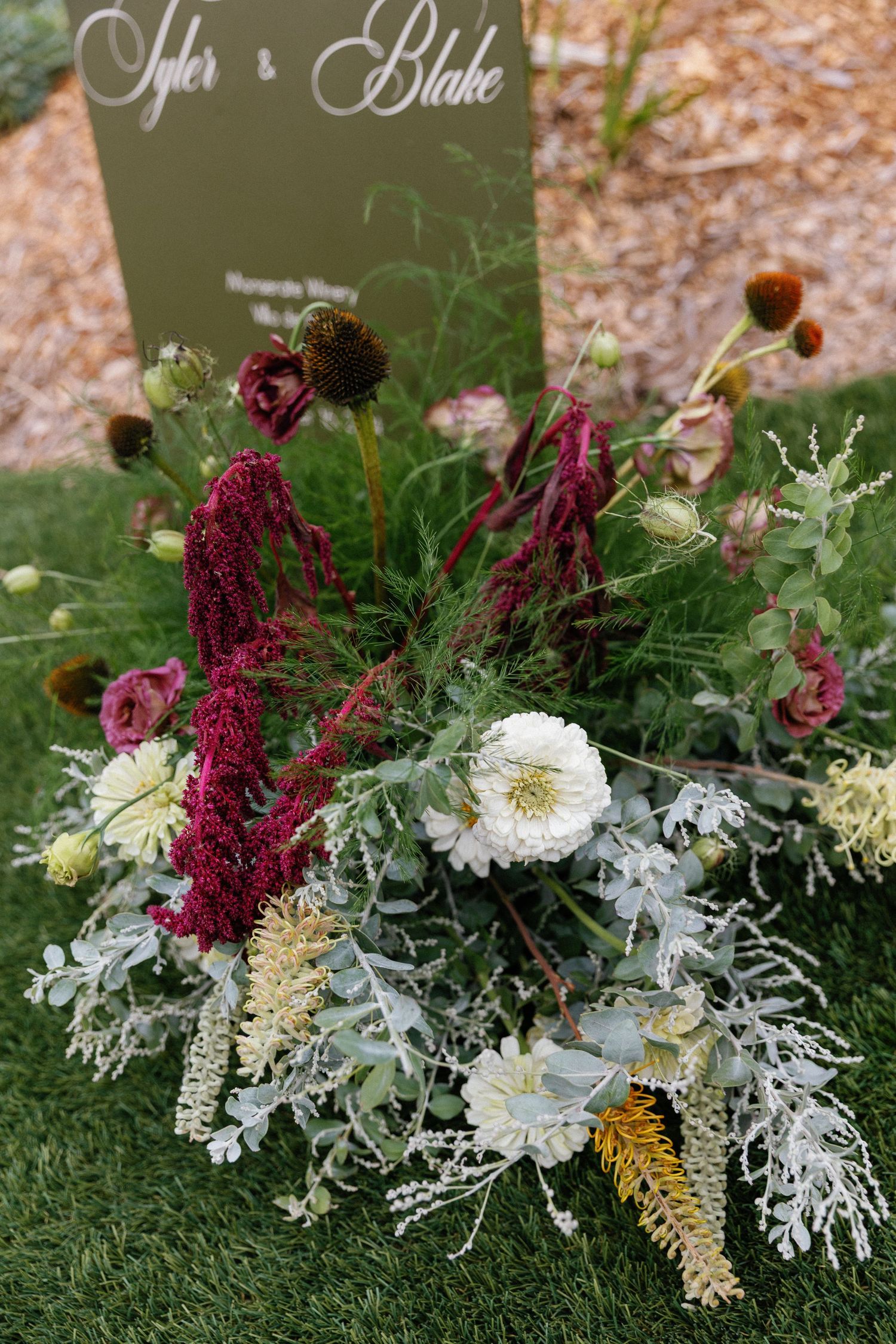 Rustic wedding floral arrangement featuring burgundy amaranthus, white daisies, and dusty miller against a sage green sign.