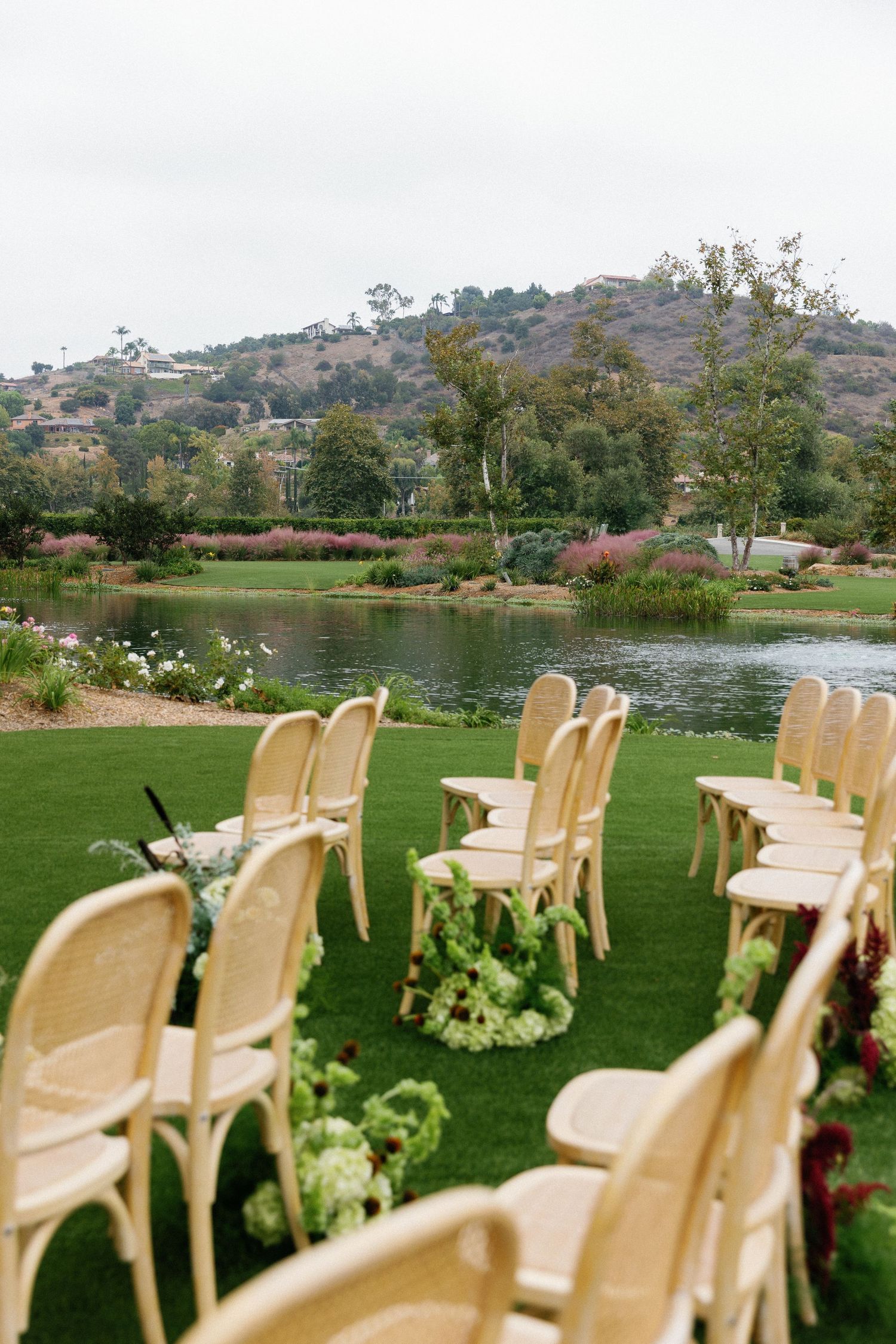 Elegant cream-colored chairs arranged for an outdoor wedding ceremony beside a scenic lake with hills in the background.