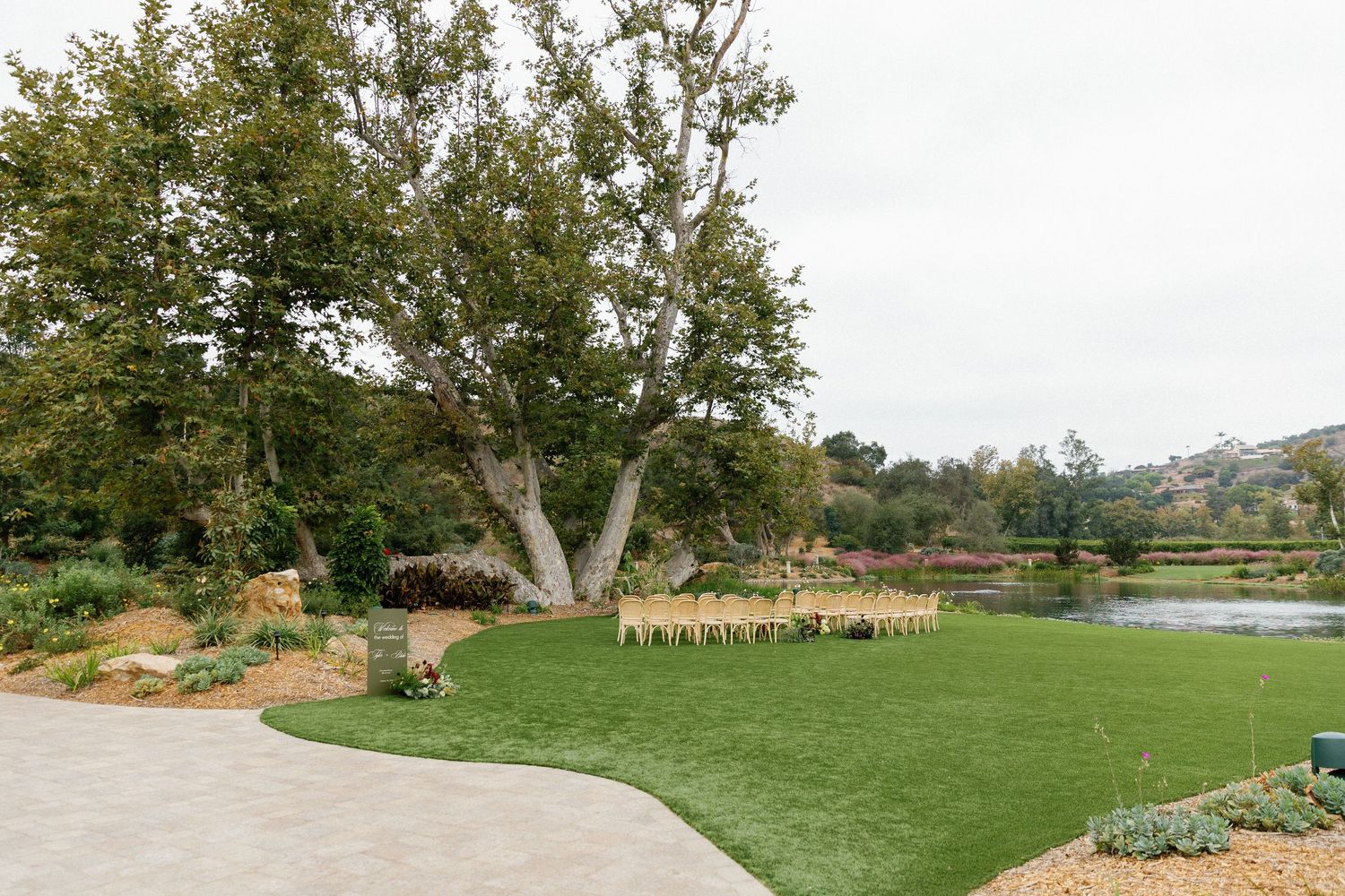 A lush garden venue with a manicured lawn, large tree, and wooden ceremony chairs set up for an outdoor wedding.