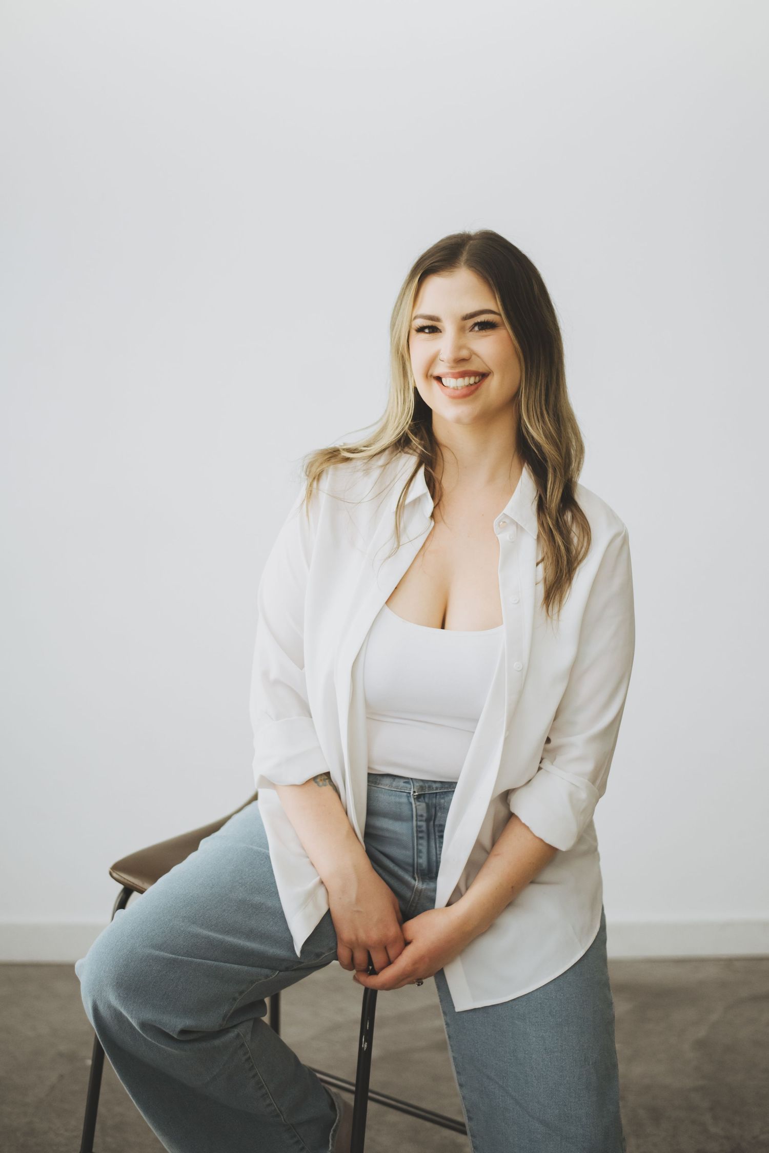Casual portrait of a woman against white wall wearing white button down shirt, tank top and jeans while sitting on stool.