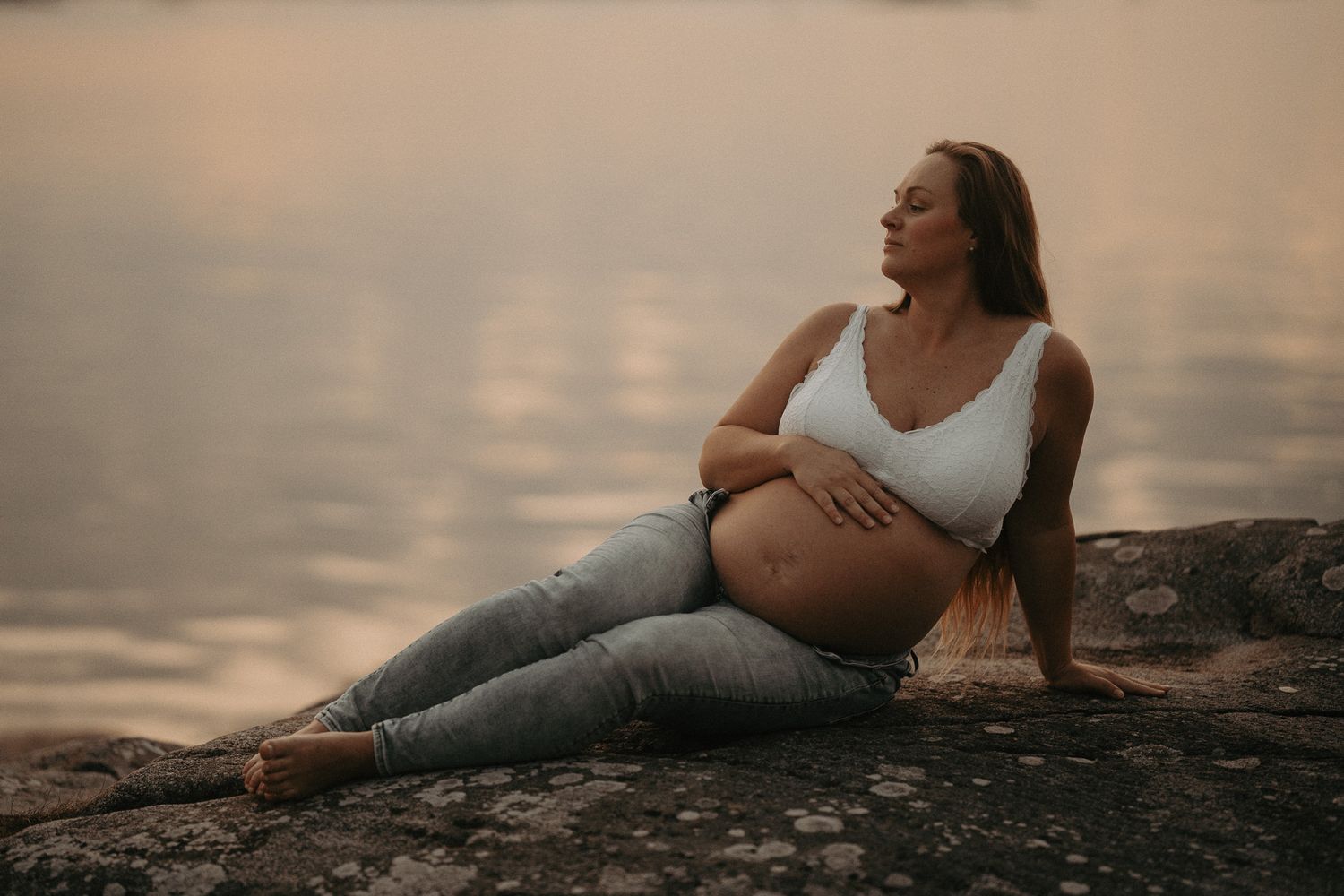 A figure reclining on rocks by water during sunset.