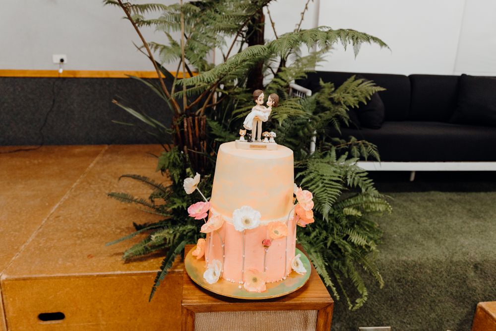 A peach-colored two-tier wedding cake decorated with flowers sits on a wooden table surrounded by ferns.