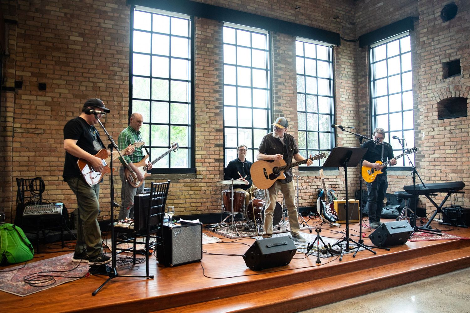 Musicians perform in a bright industrial space with brick walls and tall windows during a daytime recording session.