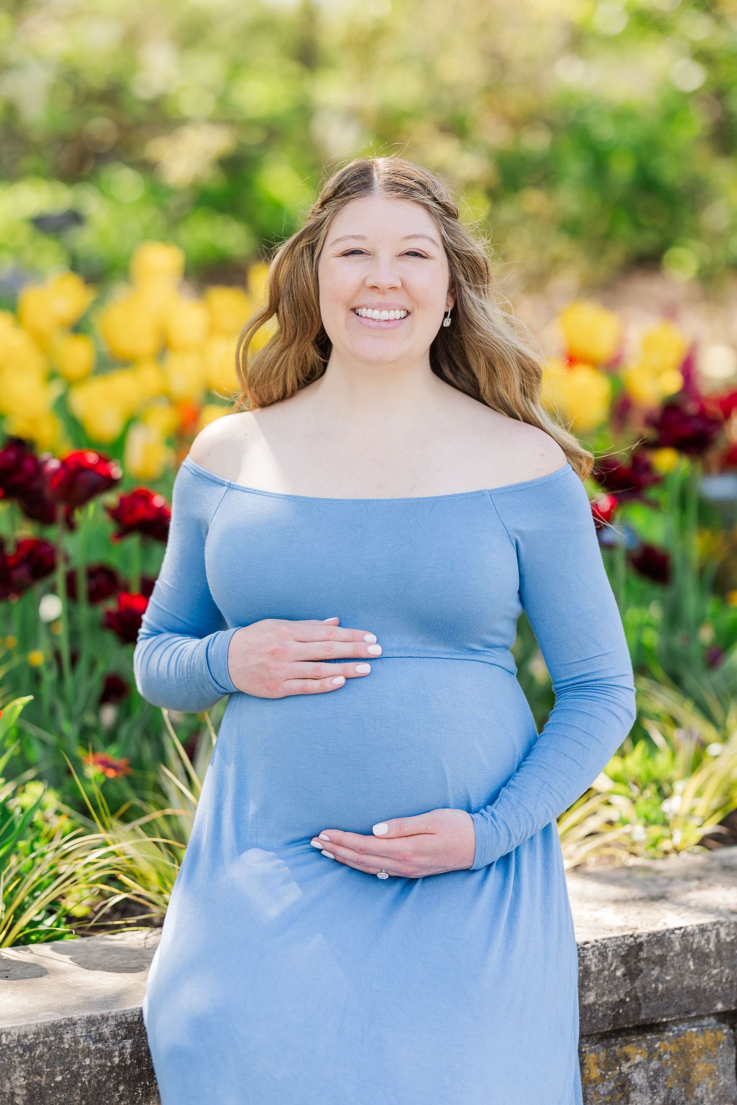 Expecting mother in light blue off-shoulder dress poses in garden with yellow and red flowers in background at Chicago Botanic Garden