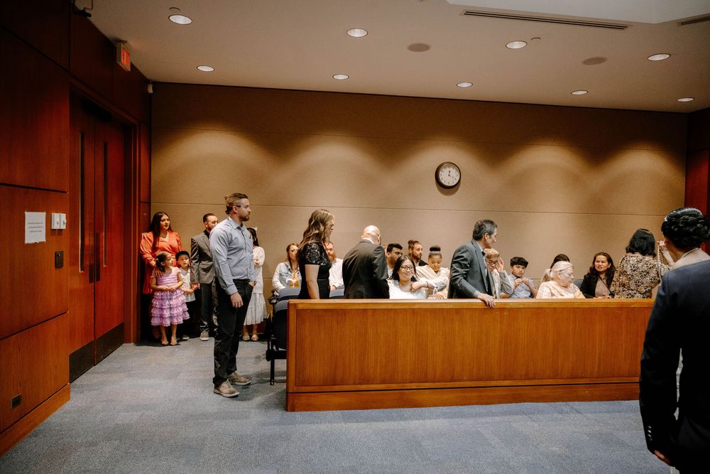 People gather in a modern courtroom with wooden paneling and recessed lighting during what appears to be a legal proceeding.